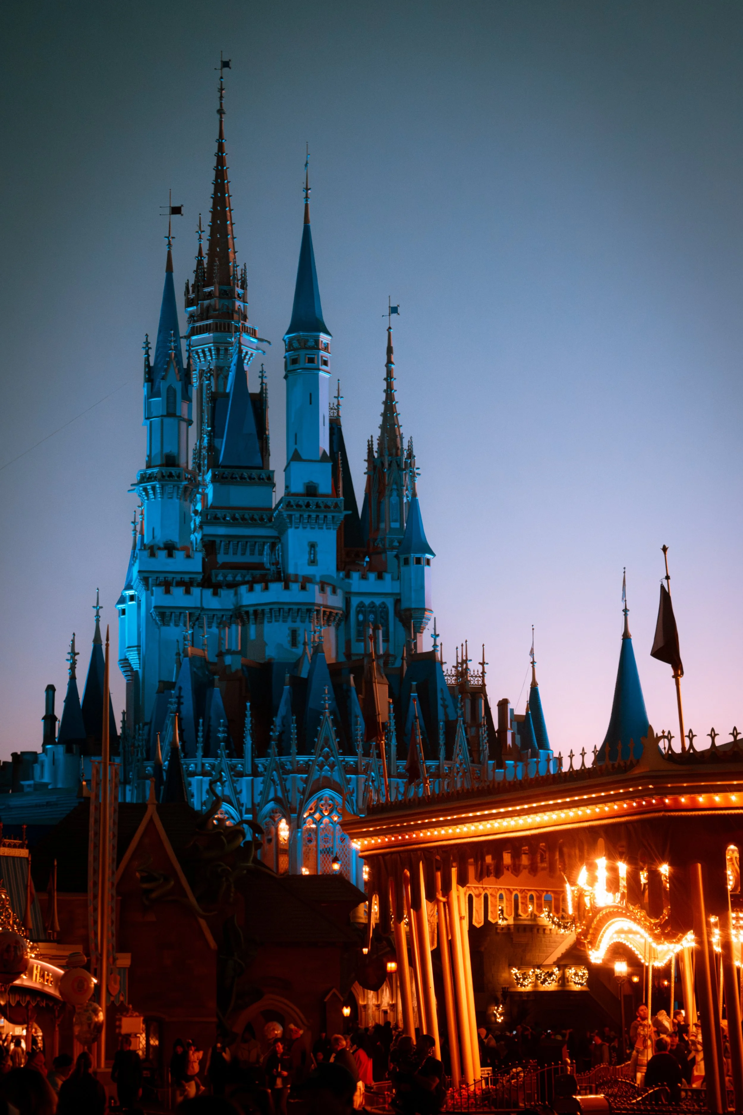 Disney castle at night with illuminated carousel in the foreground and visitors below