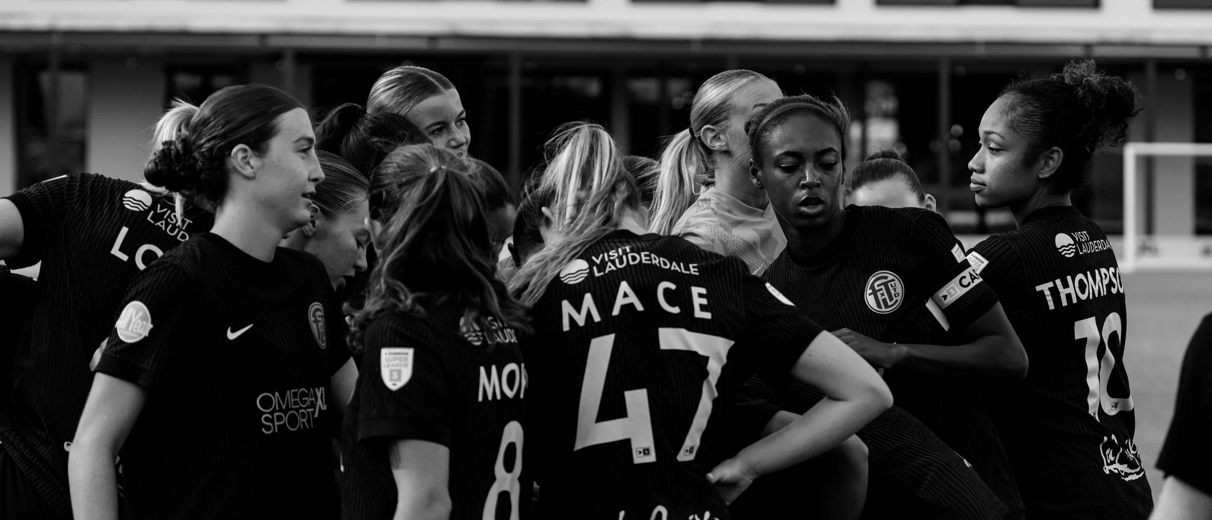 A group of female soccer players huddling together during a game, wearing black jerseys with sponsor logos and the words 'Visit Lauderdale' on them.