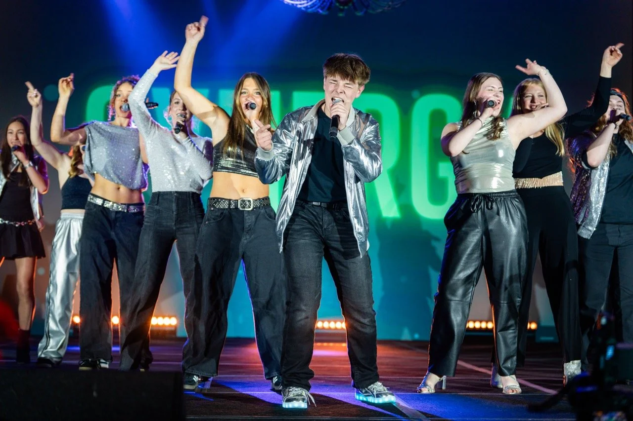 Group of young performers singing and dancing on stage during a concert or talent show, with vibrant colorful lighting in the background.