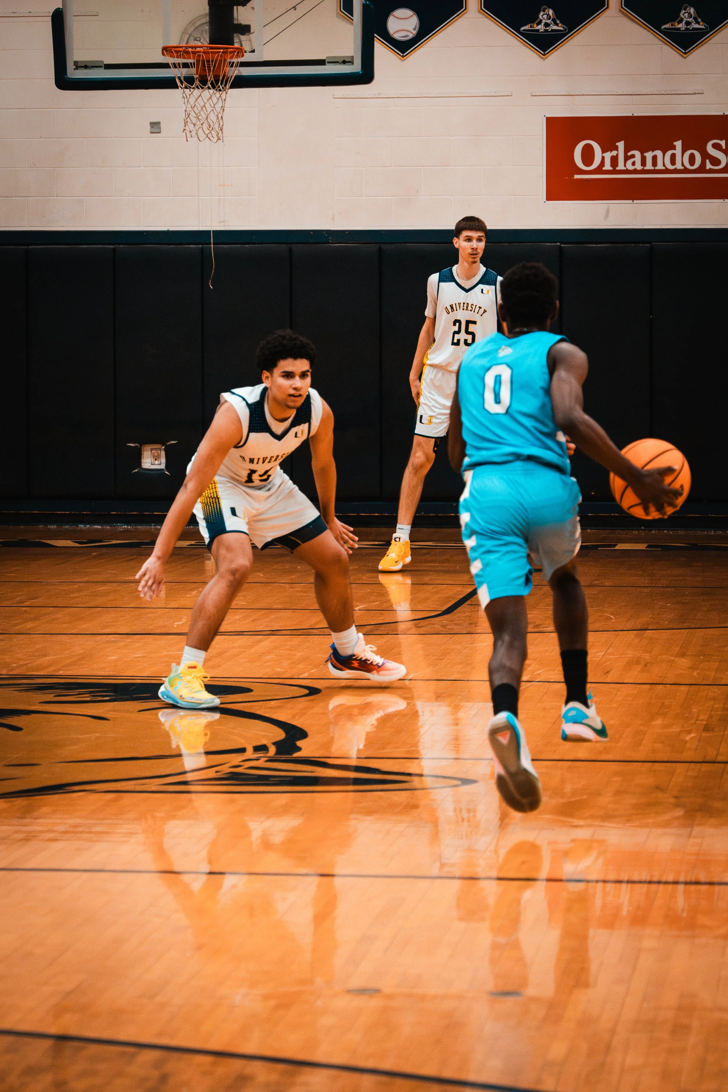 Three basketball players on an indoor court with one dribbling the ball while the others defend, with a basketball hoop above and a sign reading 'Orlando S' on the wall.