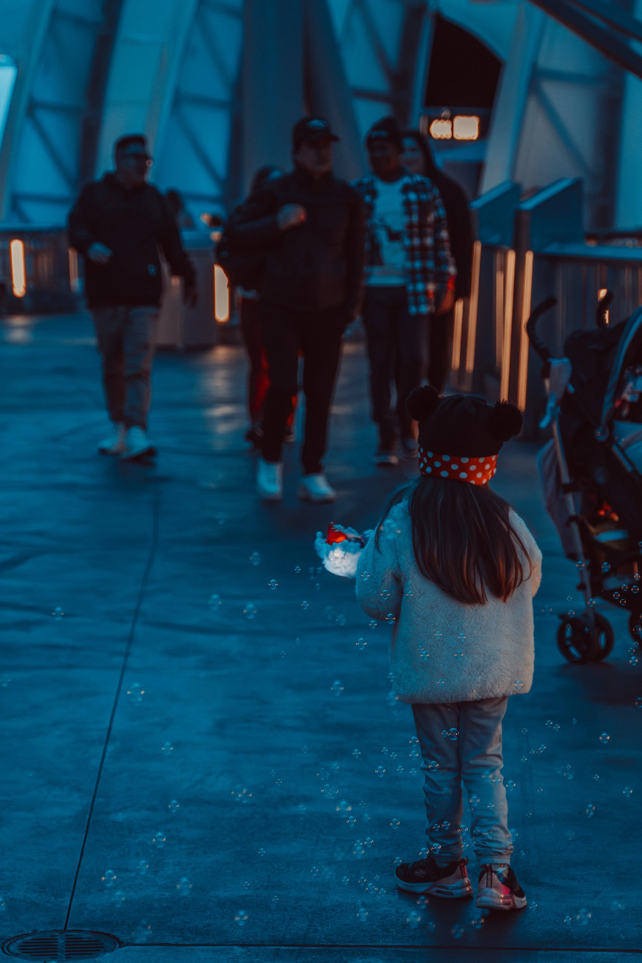 A young girl with long hair, wearing a white fuzzy jacket, polka dot headband, and sneakers, holding a bubble wand, stands on a walkway at an amusement park or similar location at dusk, with a stroller nearby and a group of people walking toward her 