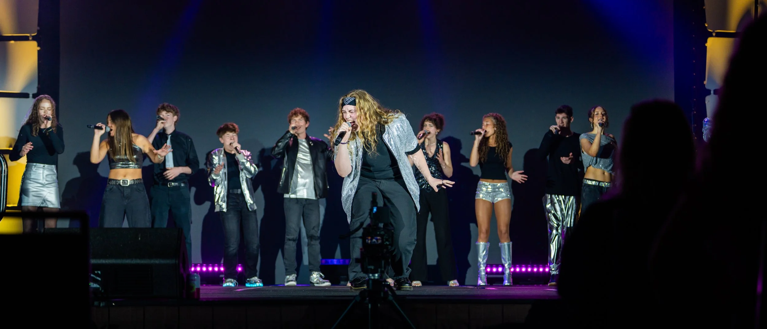 A stage with a group of young performers singing into microphones, dressed in stylish outfits, with colorful stage lighting.