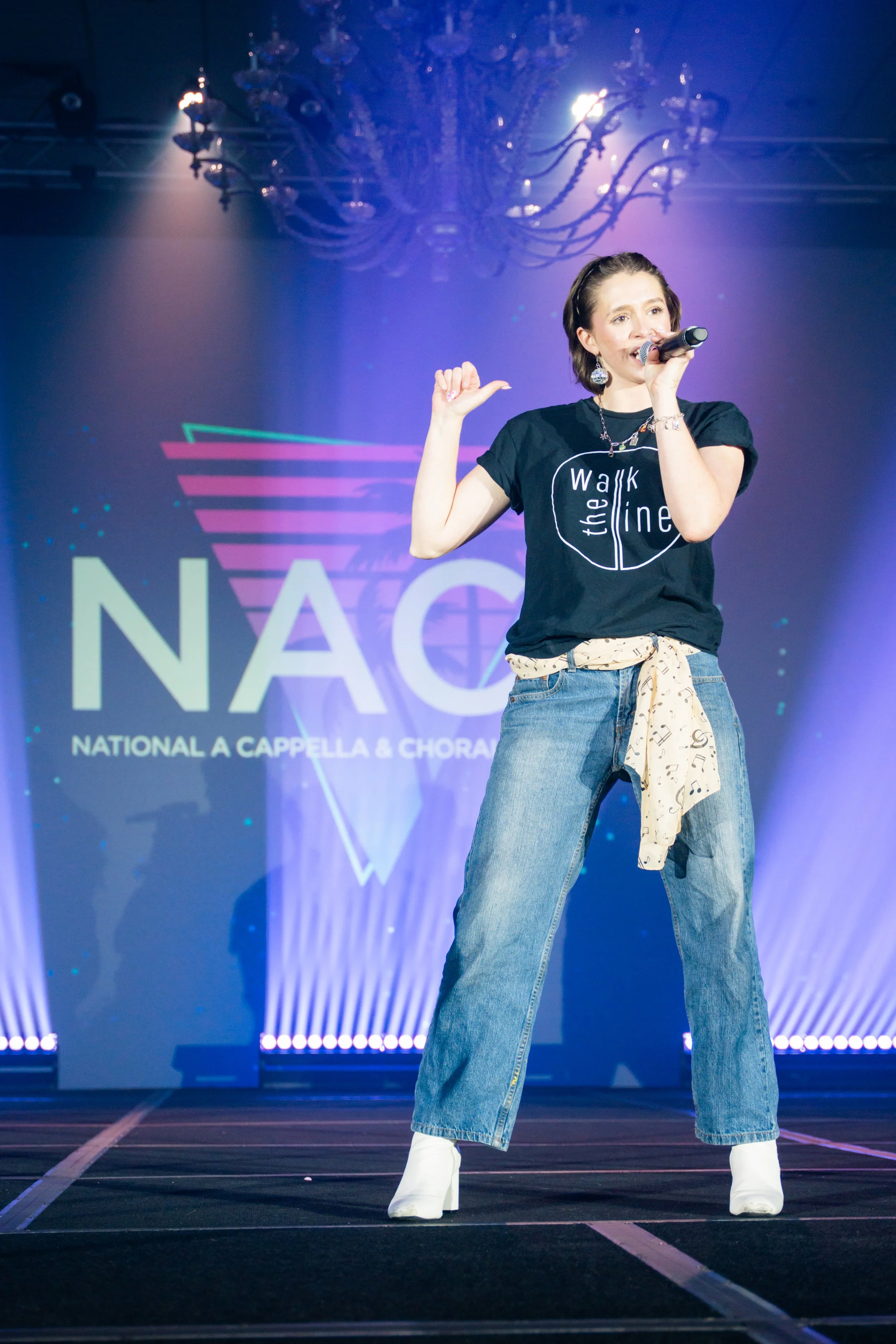 A woman performing on stage with a microphone in her right hand, wearing a black t-shirt with white text, blue jeans, white boots, and accessories, standing in front of a backdrop with the words "National A Cappella & Chorus" and colorful graphics.