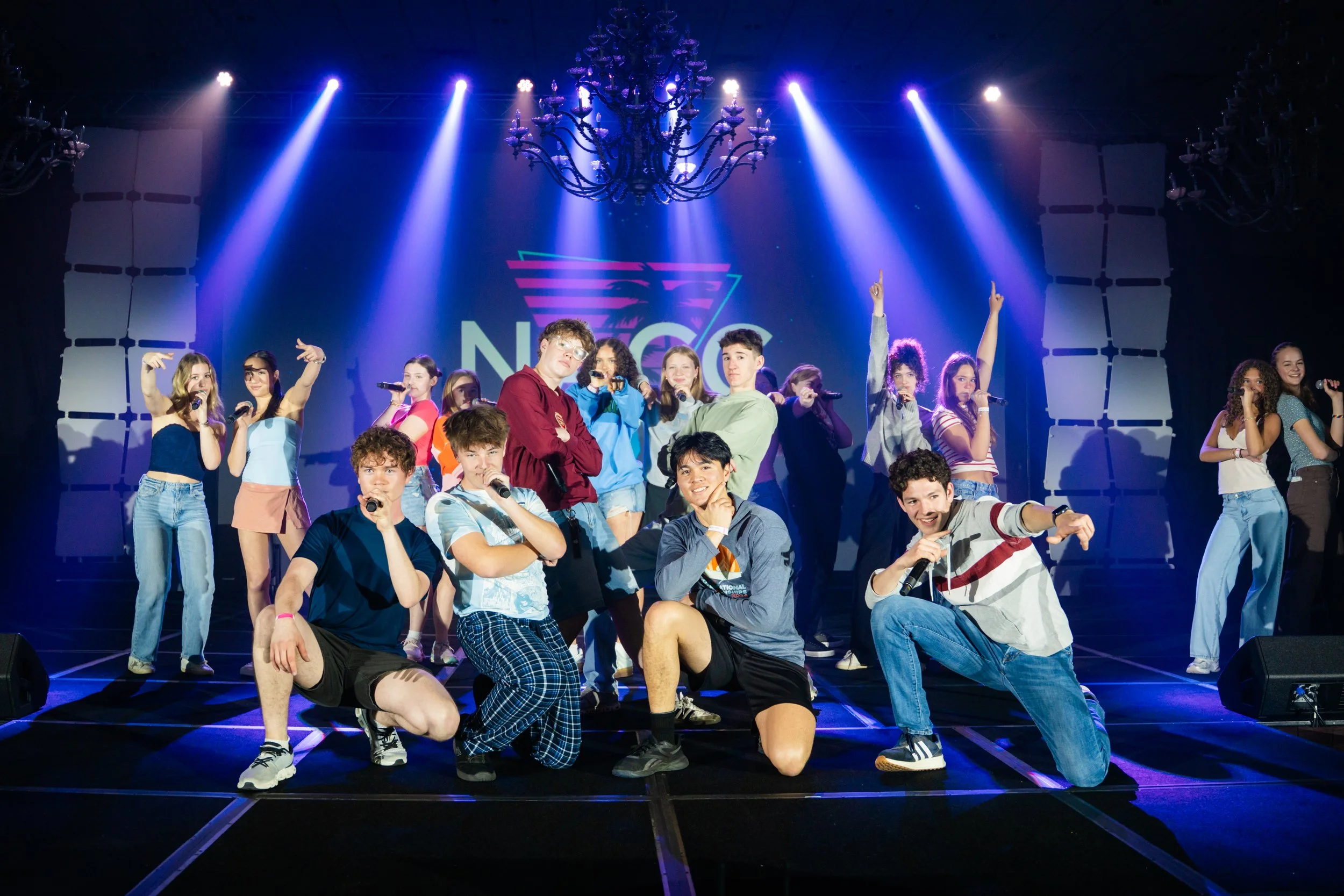 Group of young people on stage with microphones, some kneeling and some standing, under colorful stage lighting and chandeliers, with a large logo in the background that says 'NATO'.