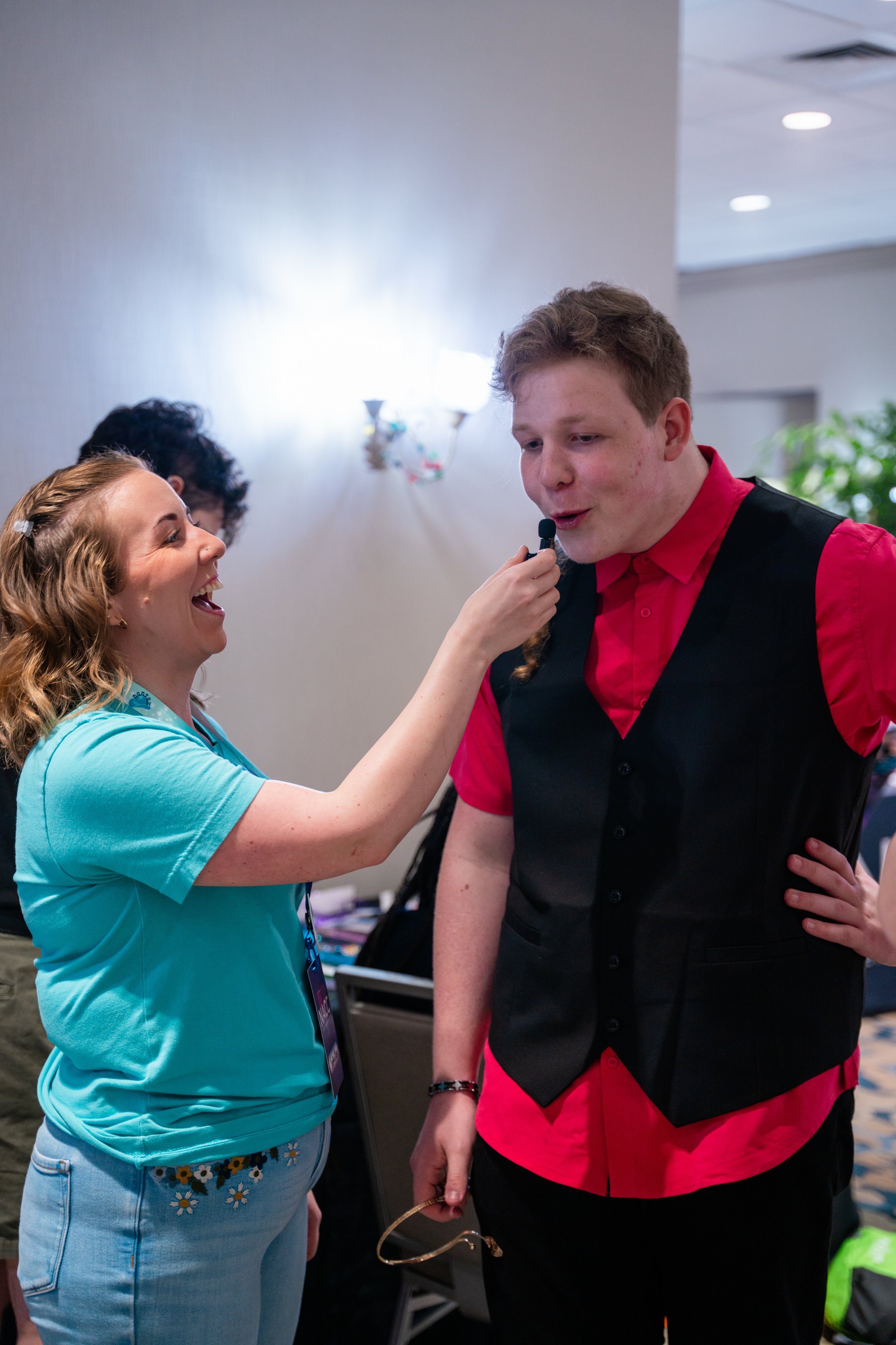 A woman in a light blue shirt and light blue jeans laughing and talking to a young man in a red shirt with a black vest, who is holding a microphone and glasses. They are indoors, with other people and a beige wall in the background.