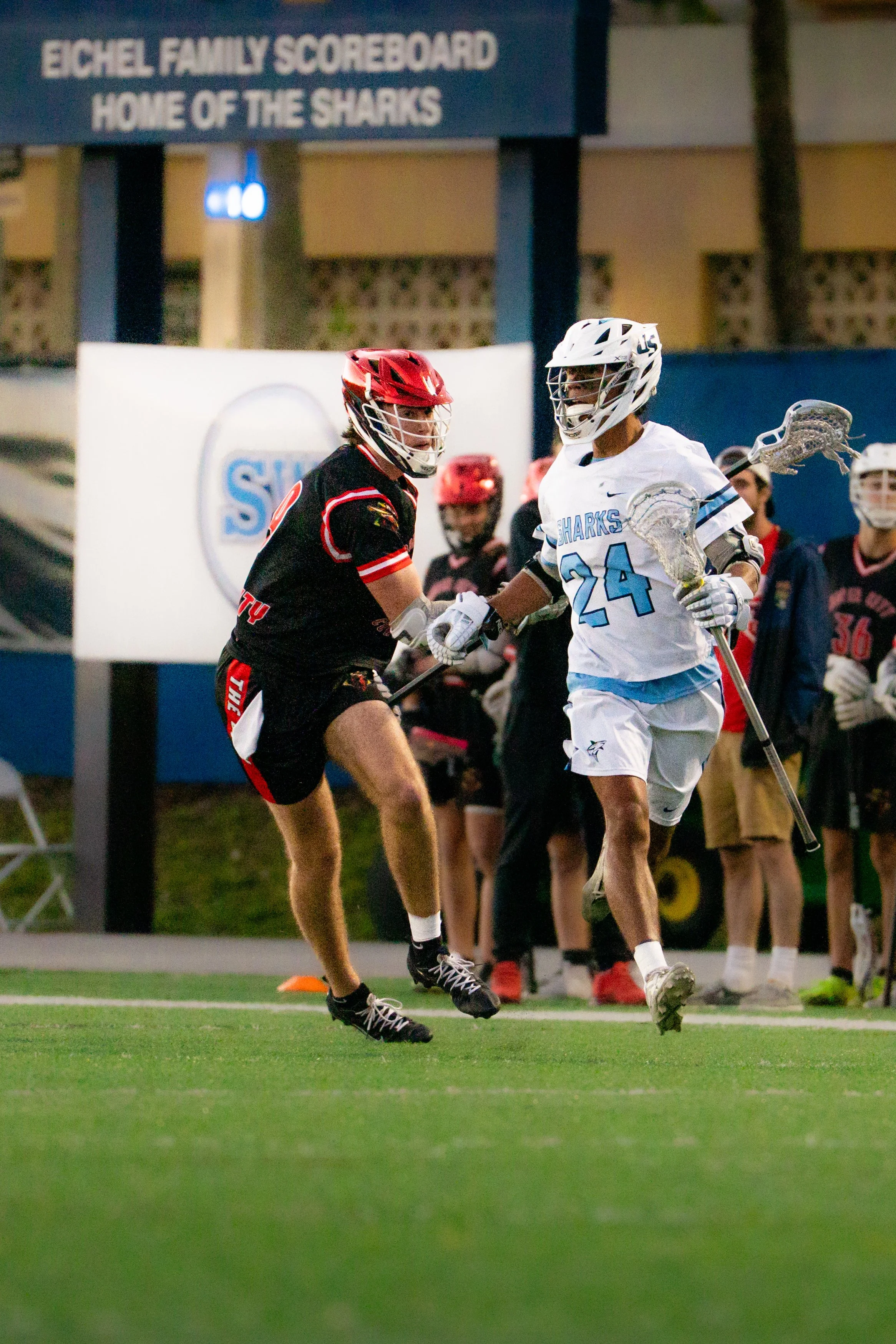 Two high school lacrosse players, one in a black and red uniform and the other in a white and light blue uniform, shake hands after a game on a green field at night. In the background, a sign reads 'Eichel Family Scoreboard' and 'Home of the Sharks'.