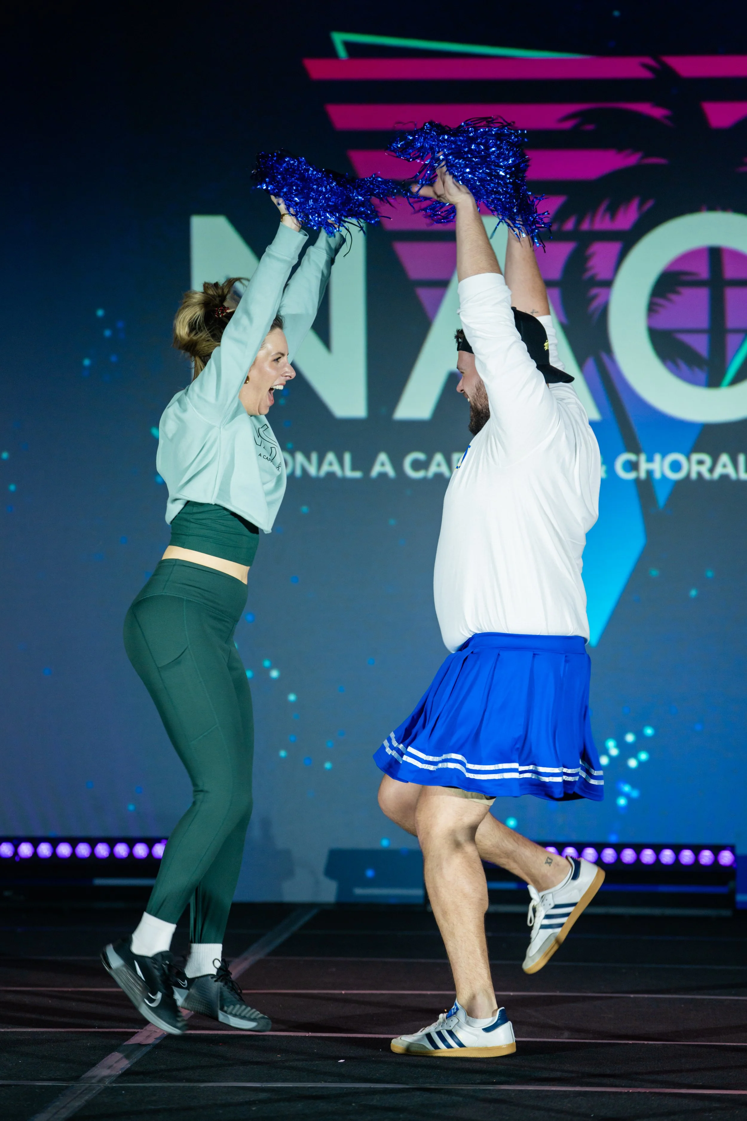 Two performers on stage, a woman in green dance attire and a man in a blue tennis skirt with a white shirt, holding blue pom-poms up in the air in a choreographed dance move.