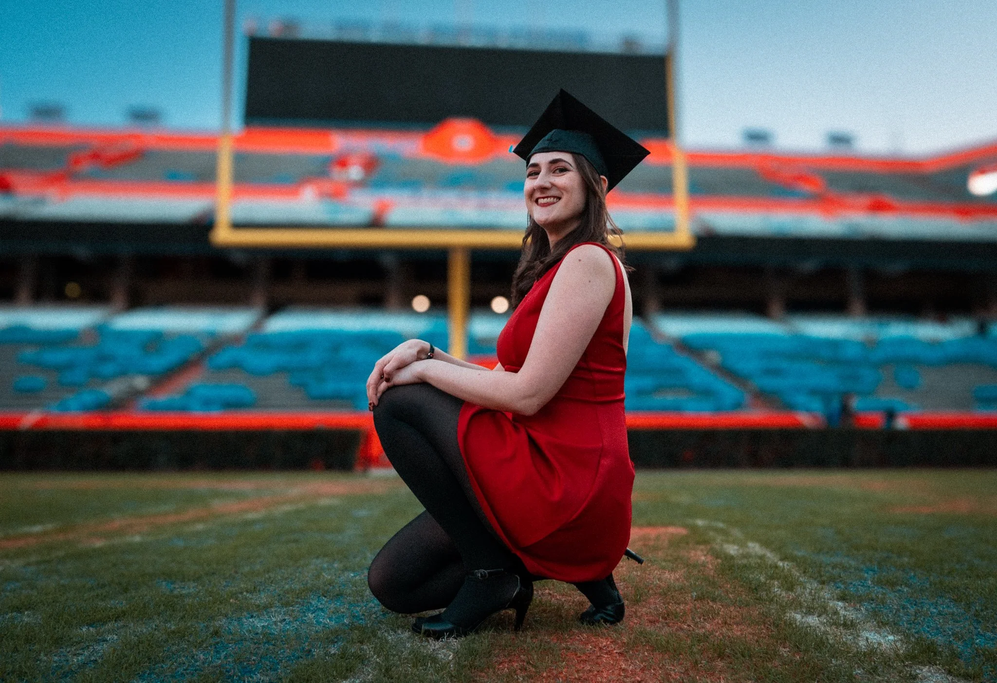 A young woman in a red dress and black high heels, wearing a graduation cap, kneeling on a football field with stadium seating in the background, smiling.