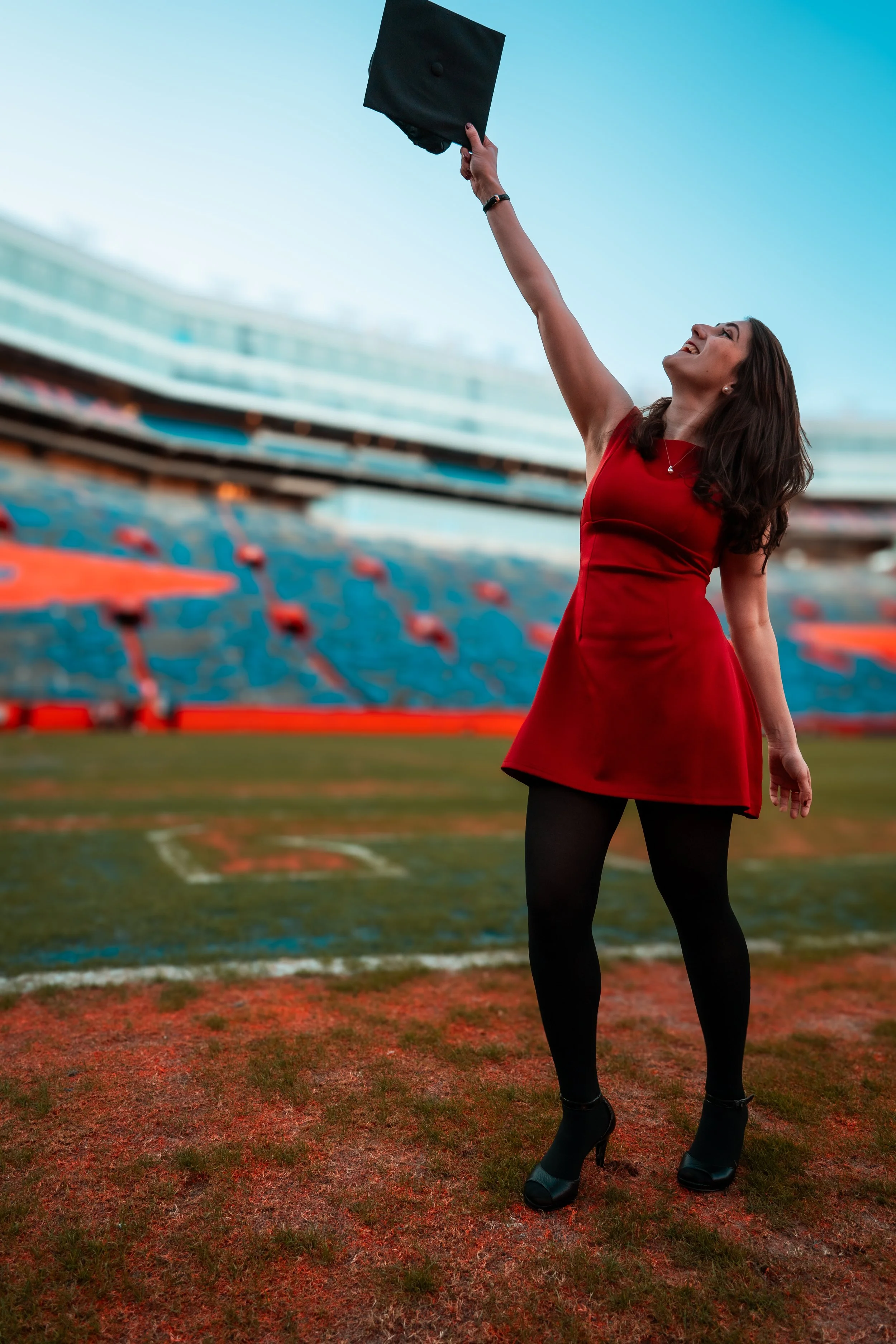A woman in a red dress standing on a sports field at a stadium, throwing a black graduation cap into the air, celebrating her graduation.
