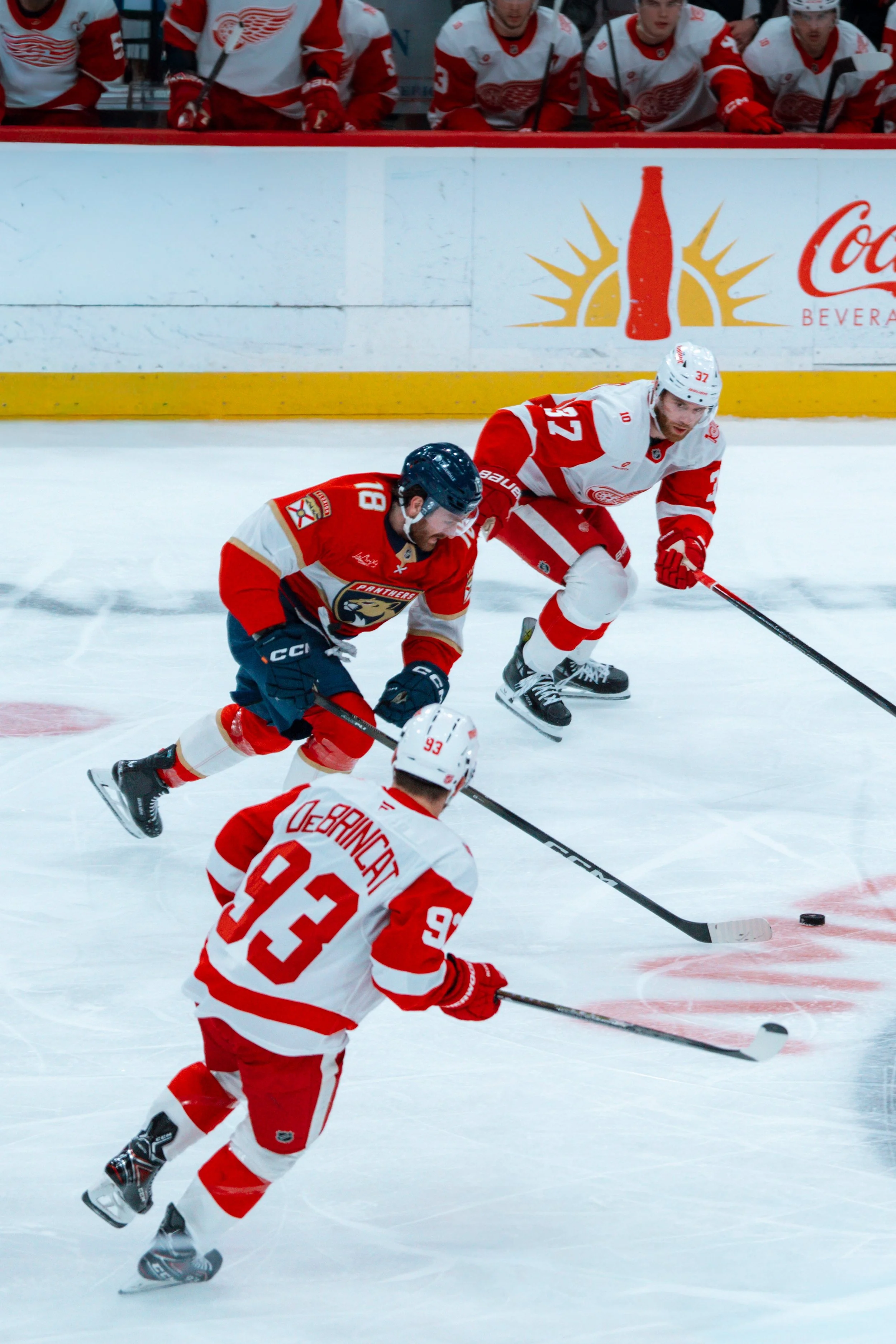 Hockey players in red and white jerseys compete for possession of the puck on ice rink with bench of players and advertising in background.