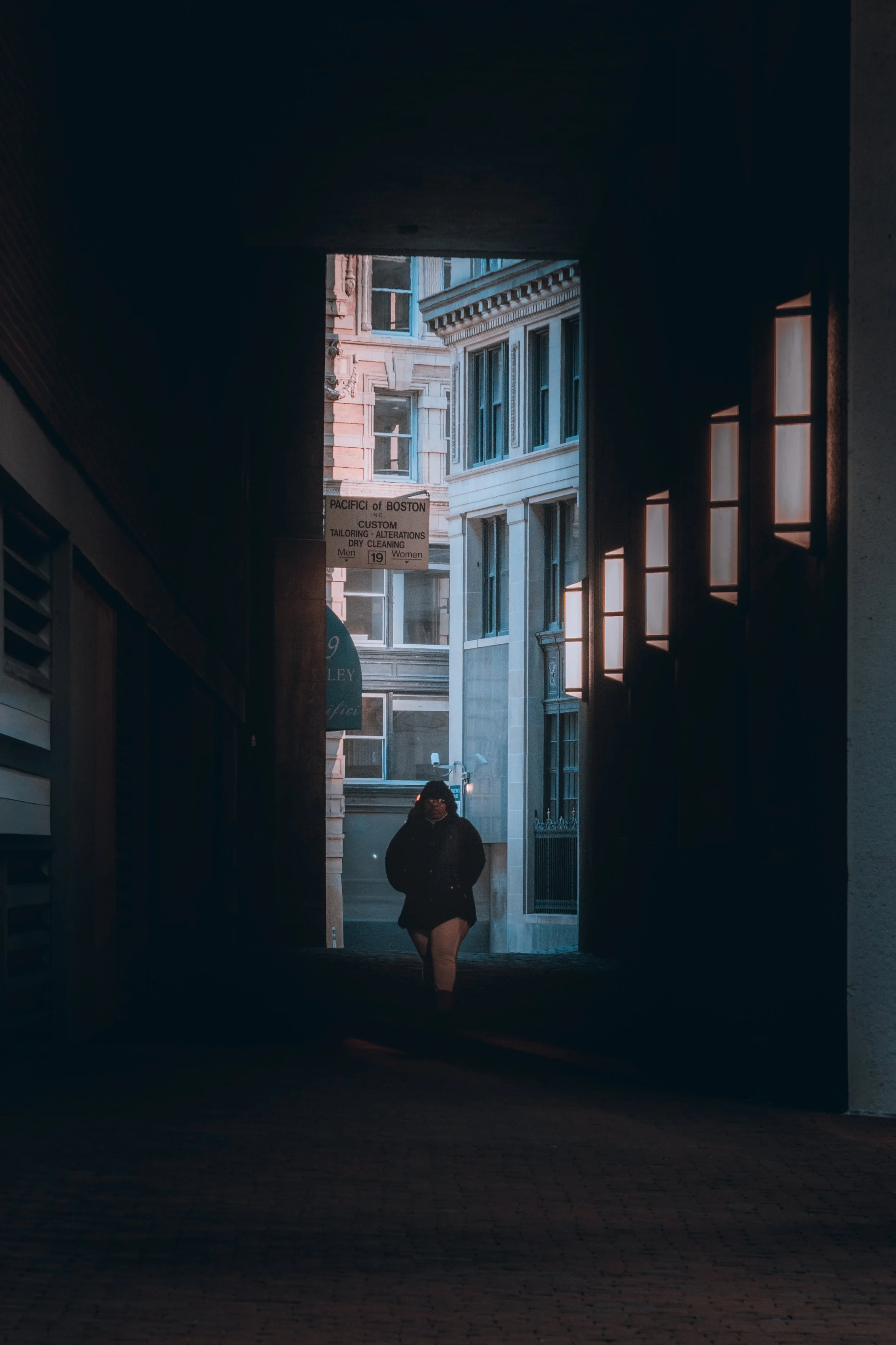 A person walking through a dark alleyway with illuminated windows on the right and a city street with buildings visible at the end of the alley.