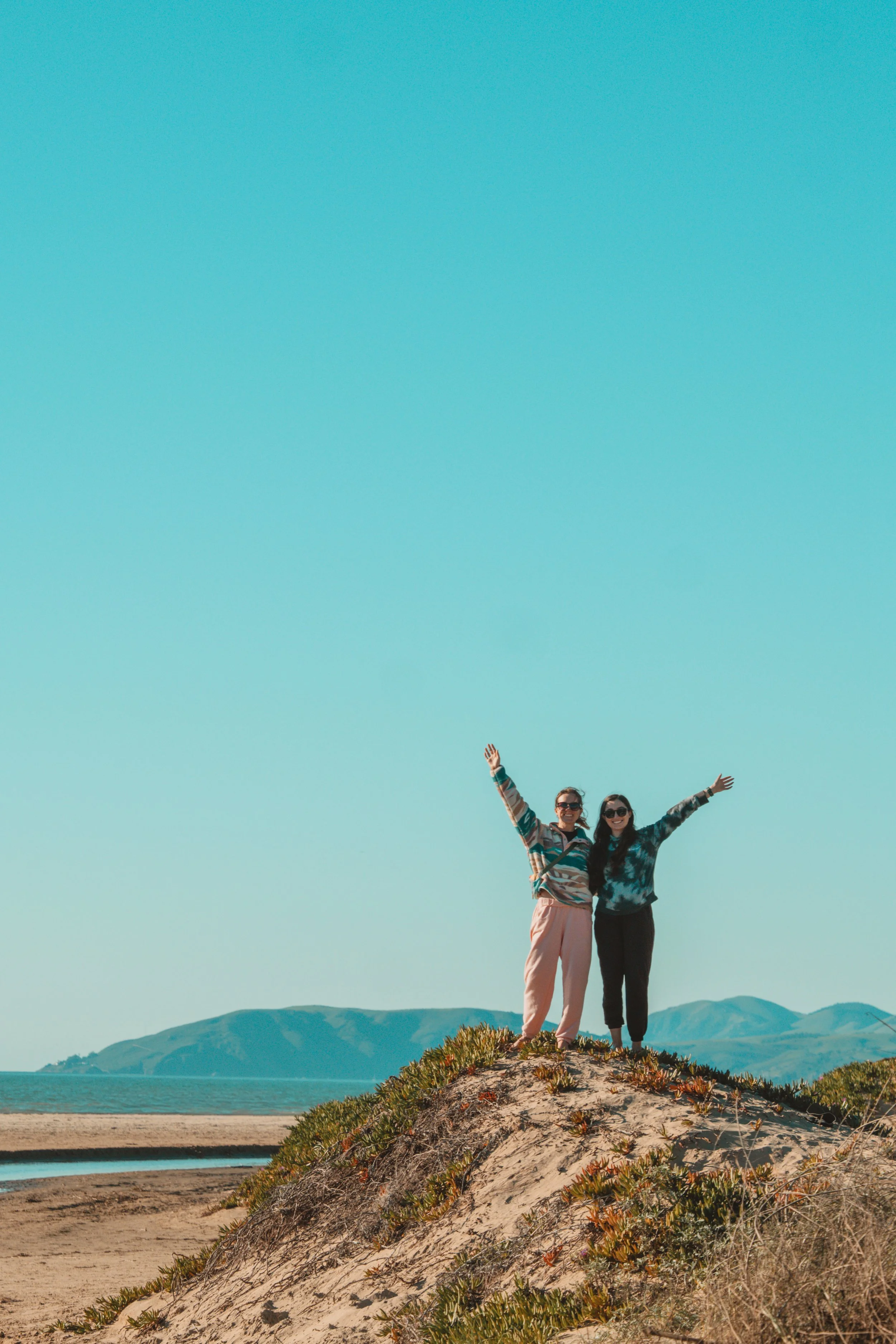 Two women standing atop a small sand hill on a beach, smiling and raising their arms in celebration, with mountains in the background under a clear blue sky.