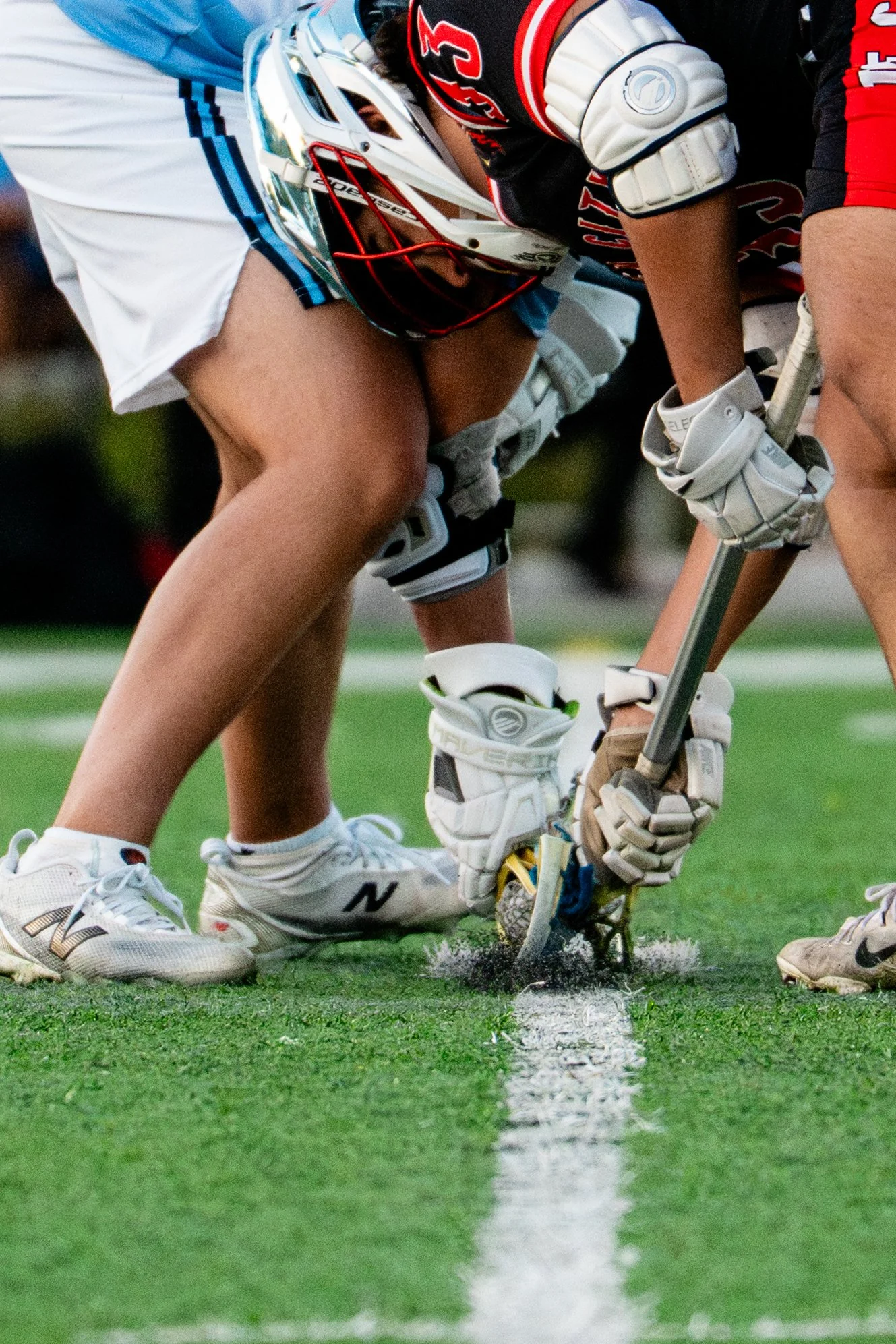 Lacrosse players crouching on the field during a game, wearing helmets and gear, with a focus on their sticks near the ground.