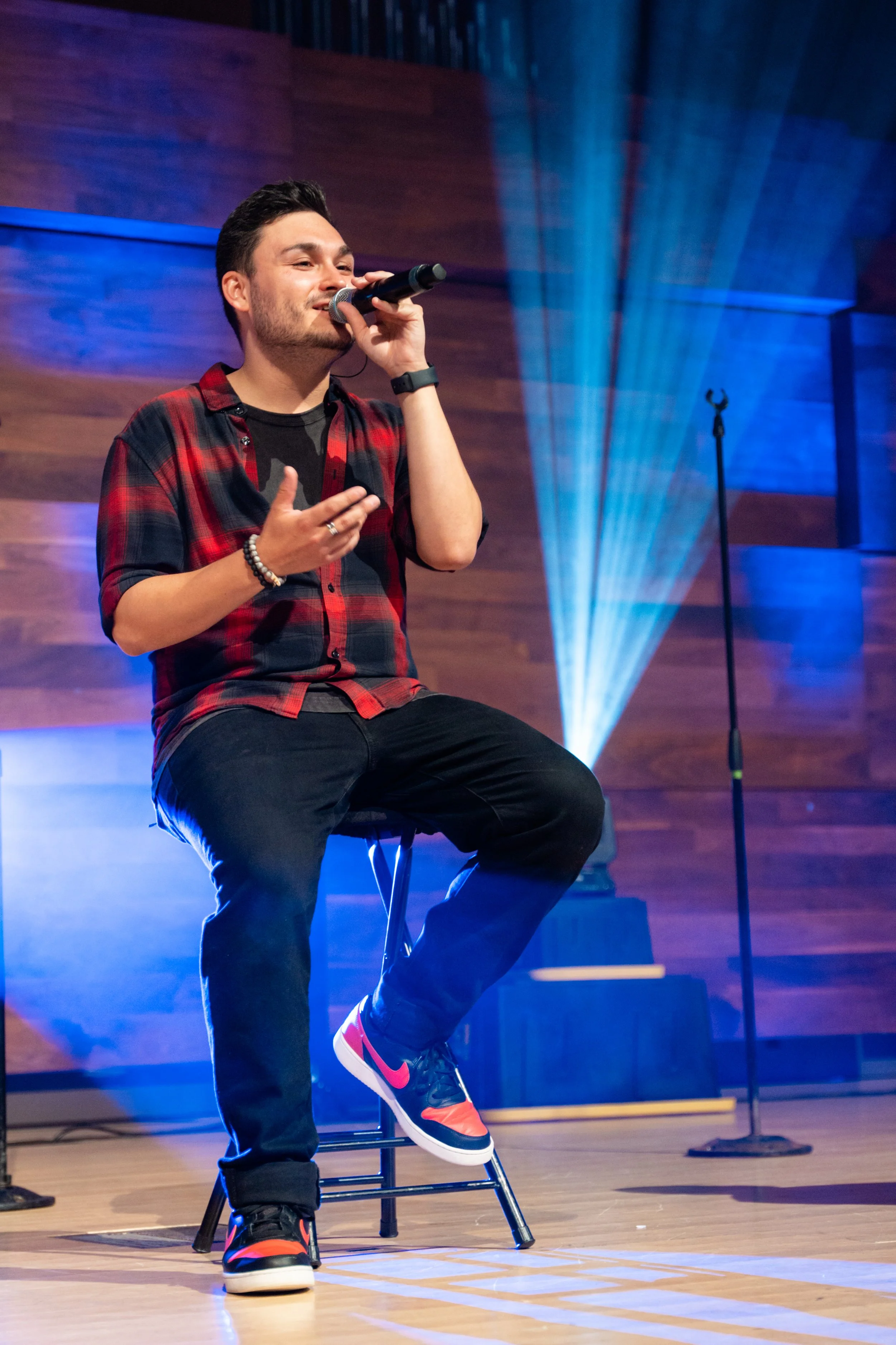 Young man sitting on a stool singing into a microphone on stage, wearing a red and black plaid shirt and sneakers, with stage lighting and a microphone stand in the background.