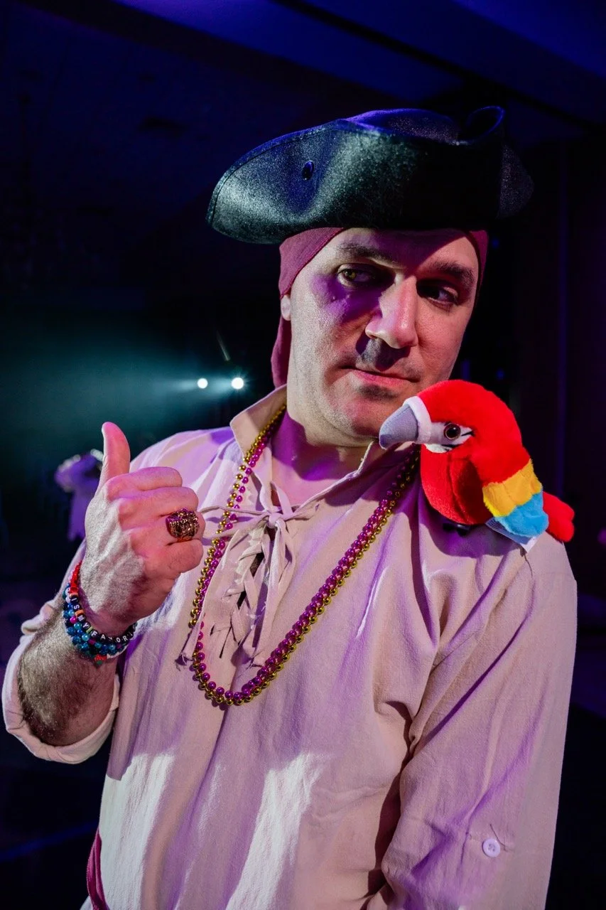 A person dressed as a pirate with a black pirate hat, maroon headscarf, and lots of beaded necklaces. They have a plush toy parrot on their shoulder and are giving a thumbs-up. The background is dark with stage lighting.