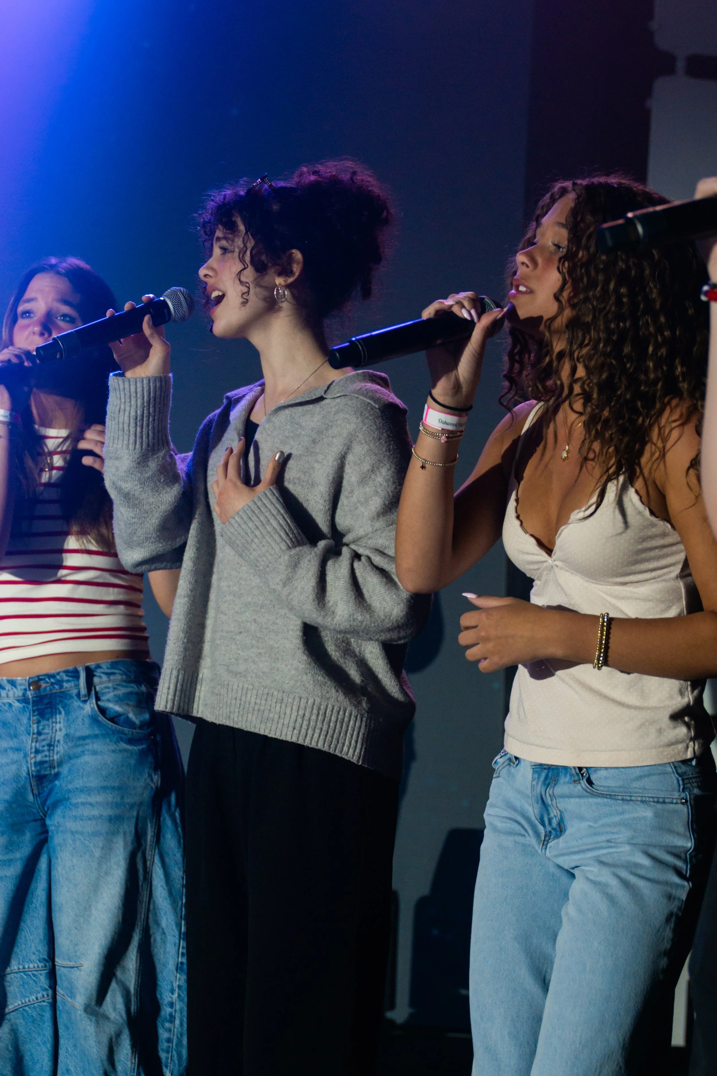 Four women singing into microphones on a stage, wearing casual clothing including jeans and tank tops.
