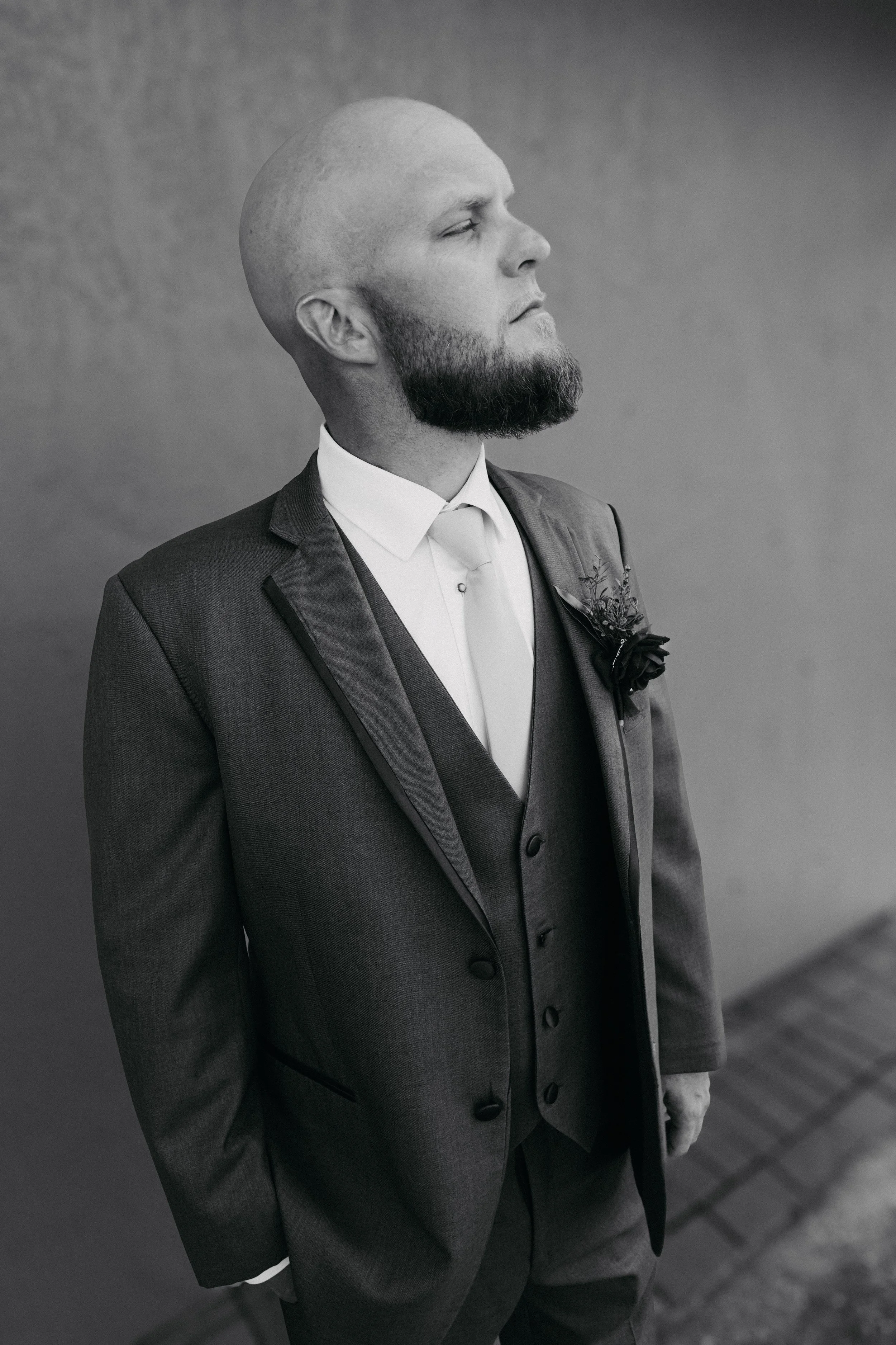 Man in formal suit with a boutonniere, standing outdoors with a contemplative expression, facing left.