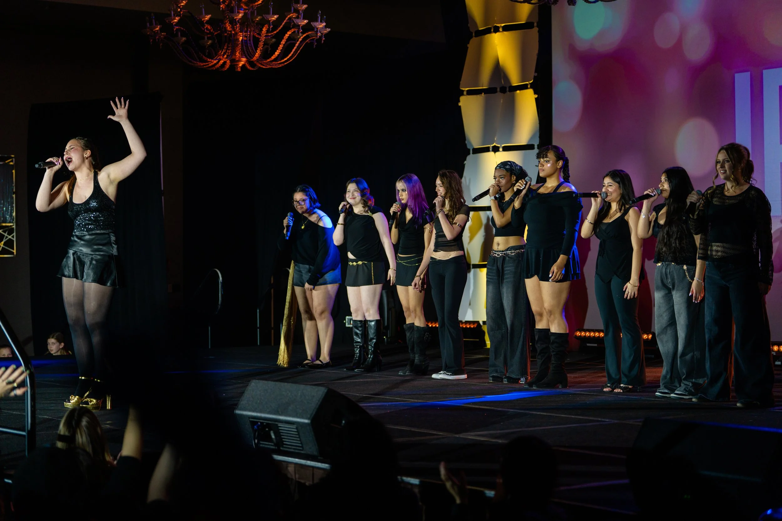 A group of young women singing and holding microphones on a stage during a performance, with one woman leading and others singing along, with stage lighting and a projection screen in the background.