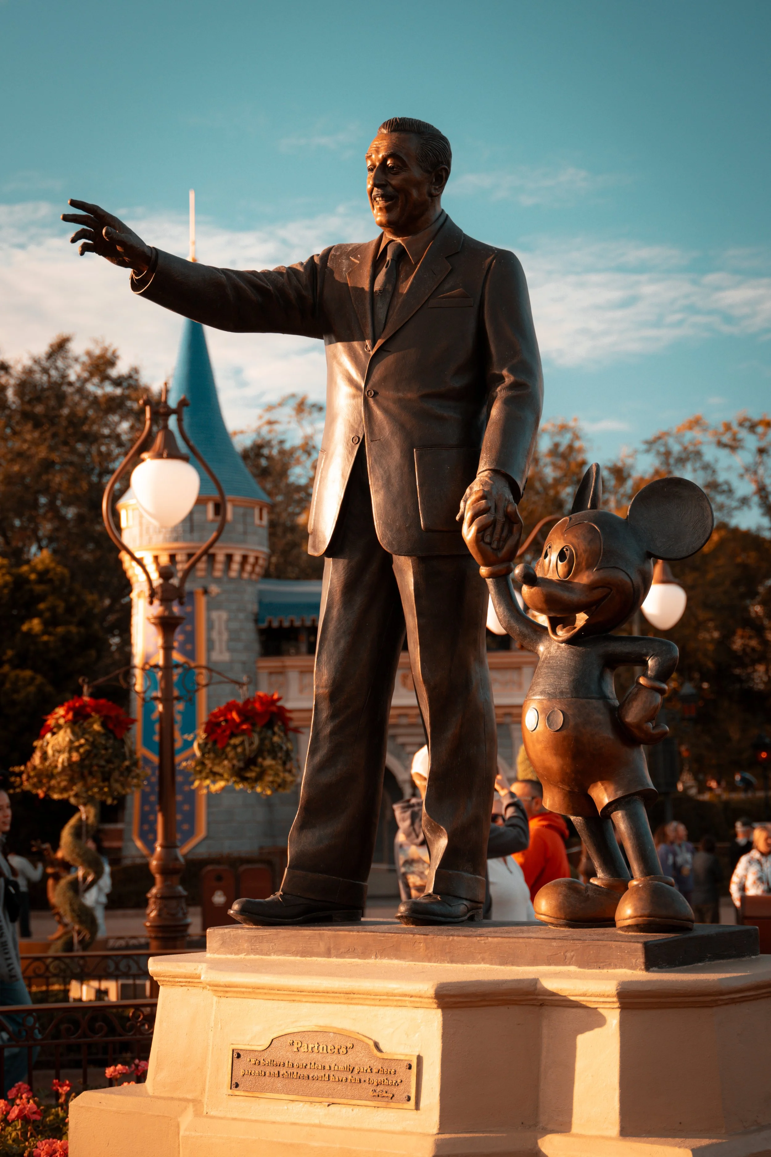 Bronze statue of Walt Disney holding hands with Mickey Mouse at Disneyland with a castle in the background during sunset.