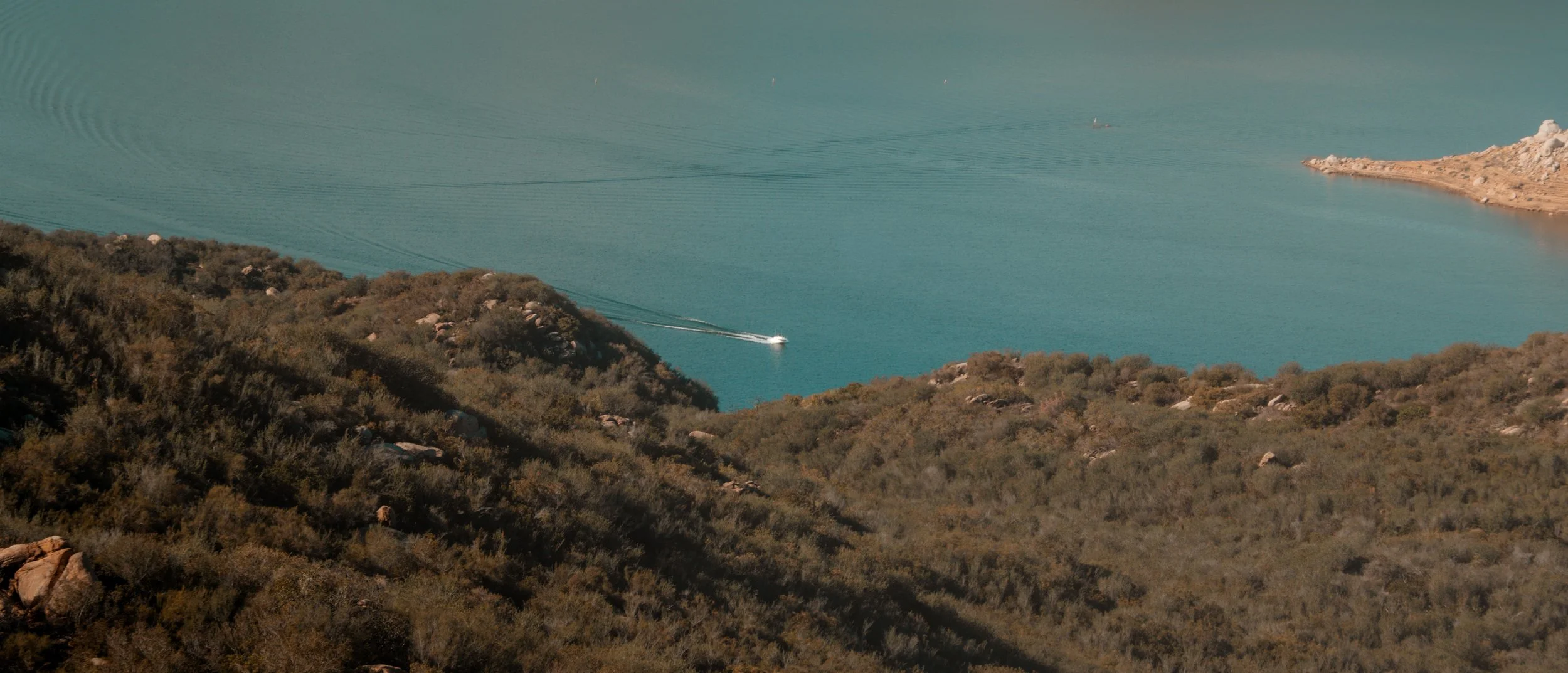 A boat sailing on a body of water with a wake trail, surrounded by green hills and rocky terrain.
