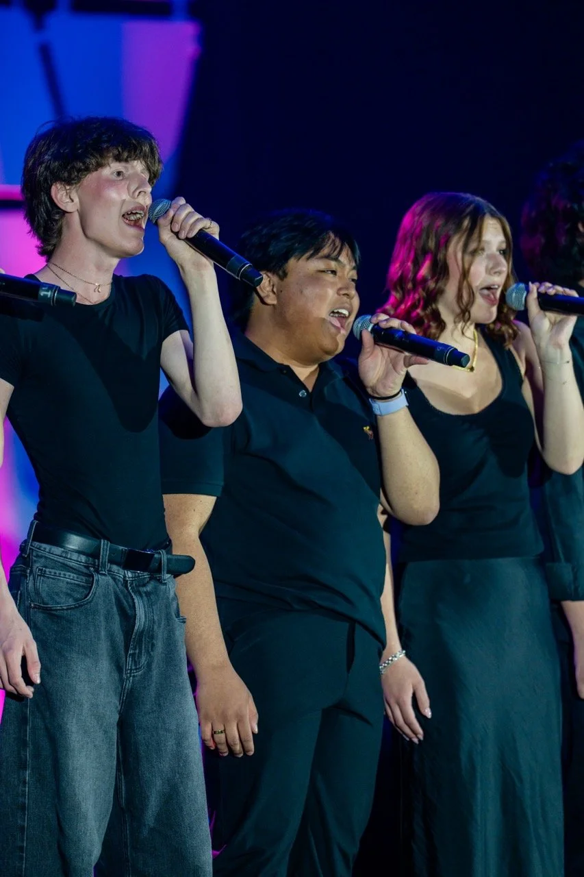 Three young people singing into microphones on stage with colorful lighting.