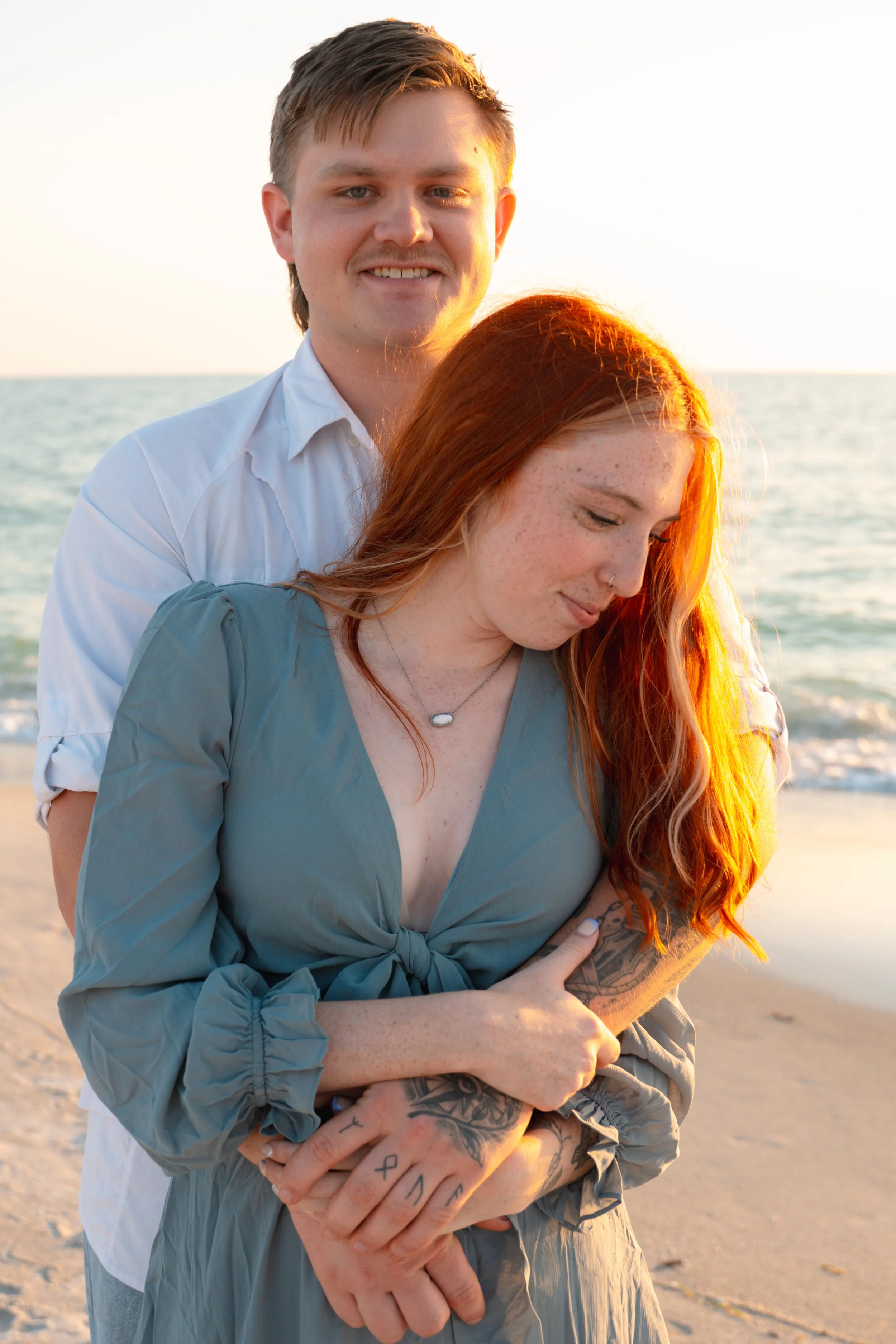 A young couple standing on the beach during sunset, with the man in a white shirt behind the woman with red hair and tattoos, both smiling and embracing.