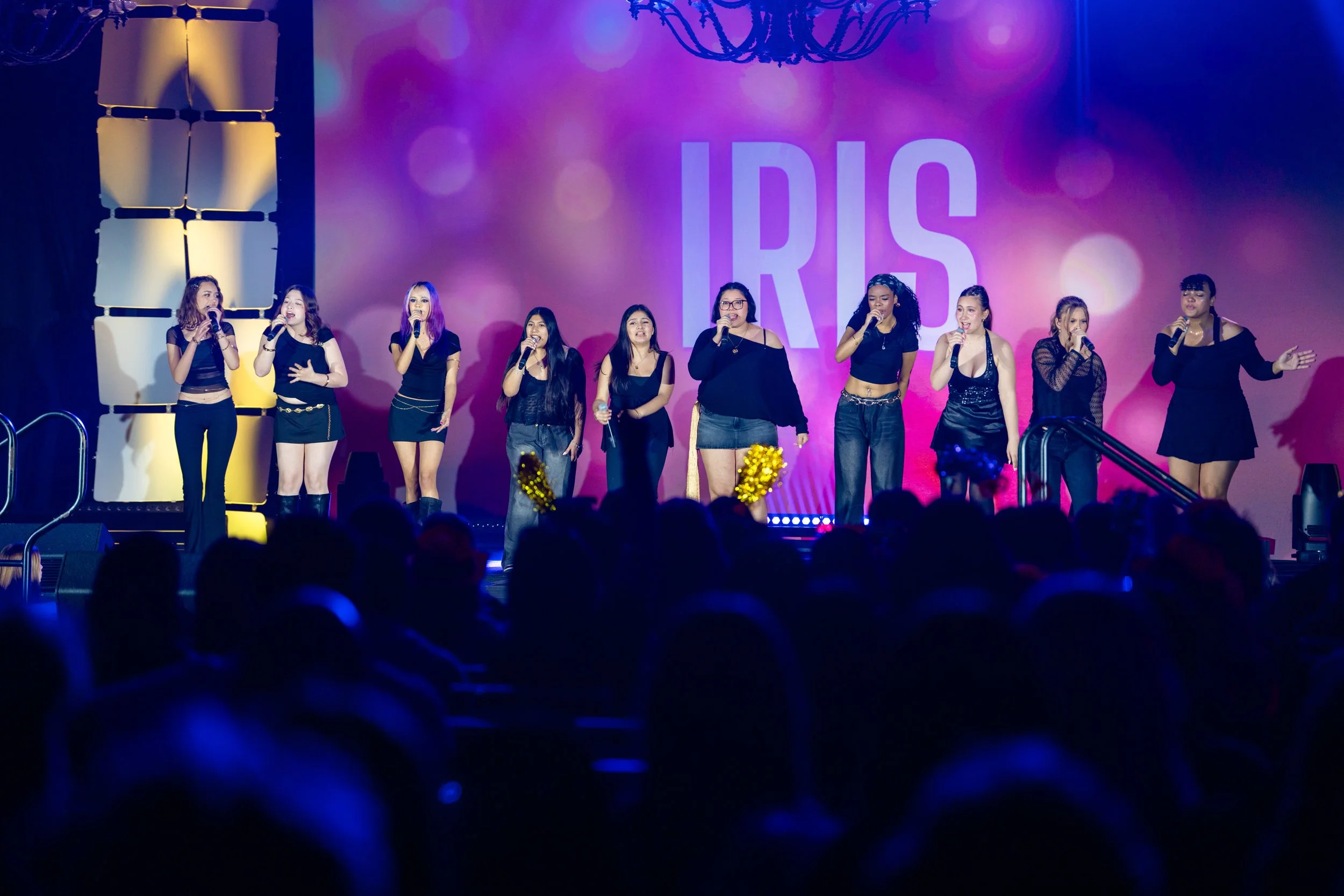 A group of female performers singing on stage at an event called 'IRIS.' The stage has colorful lighting and a large screen with the event name in the background. The audience is visible in the foreground.