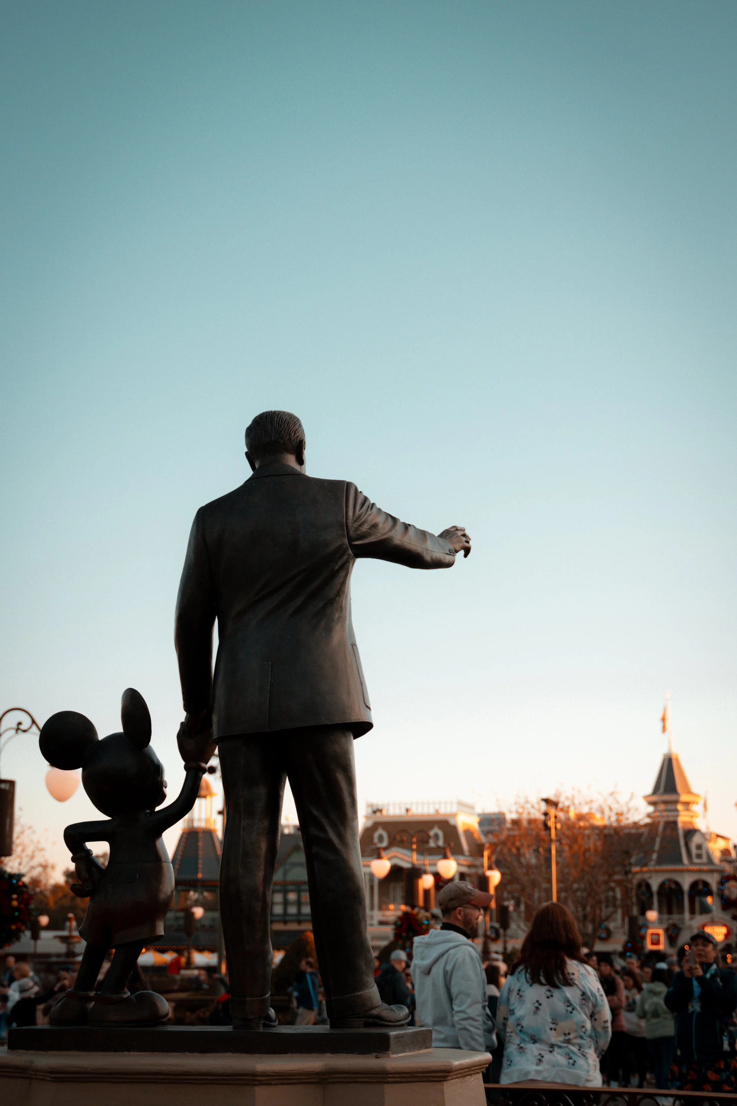 Bronze statue of Walt Disney and Minnie Mouse at Disneyland with people in the background near a Victorian-style building.