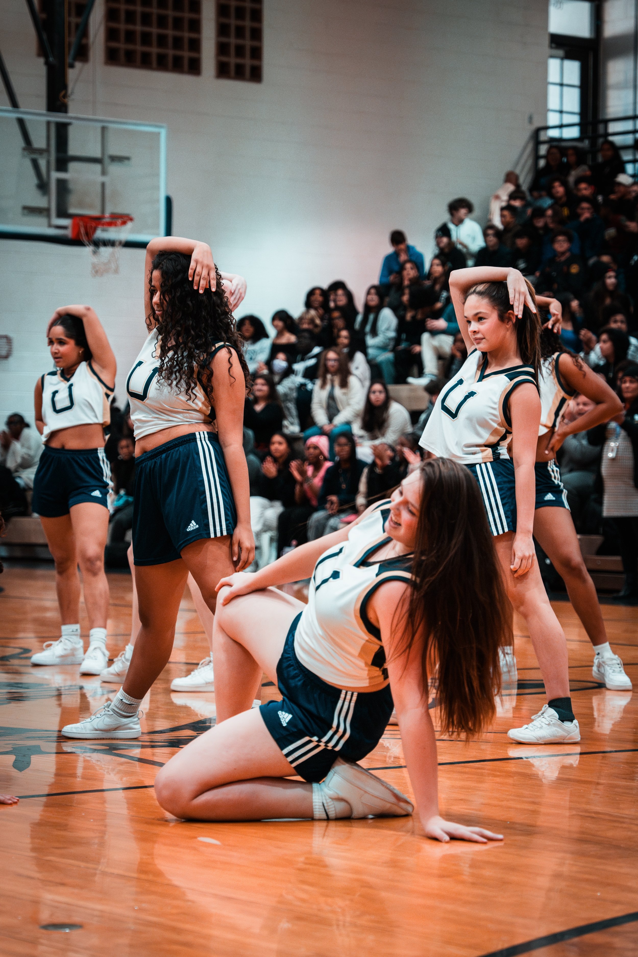Girls in cheerleading uniforms performing a dance routine on a basketball court with an audience in the background.