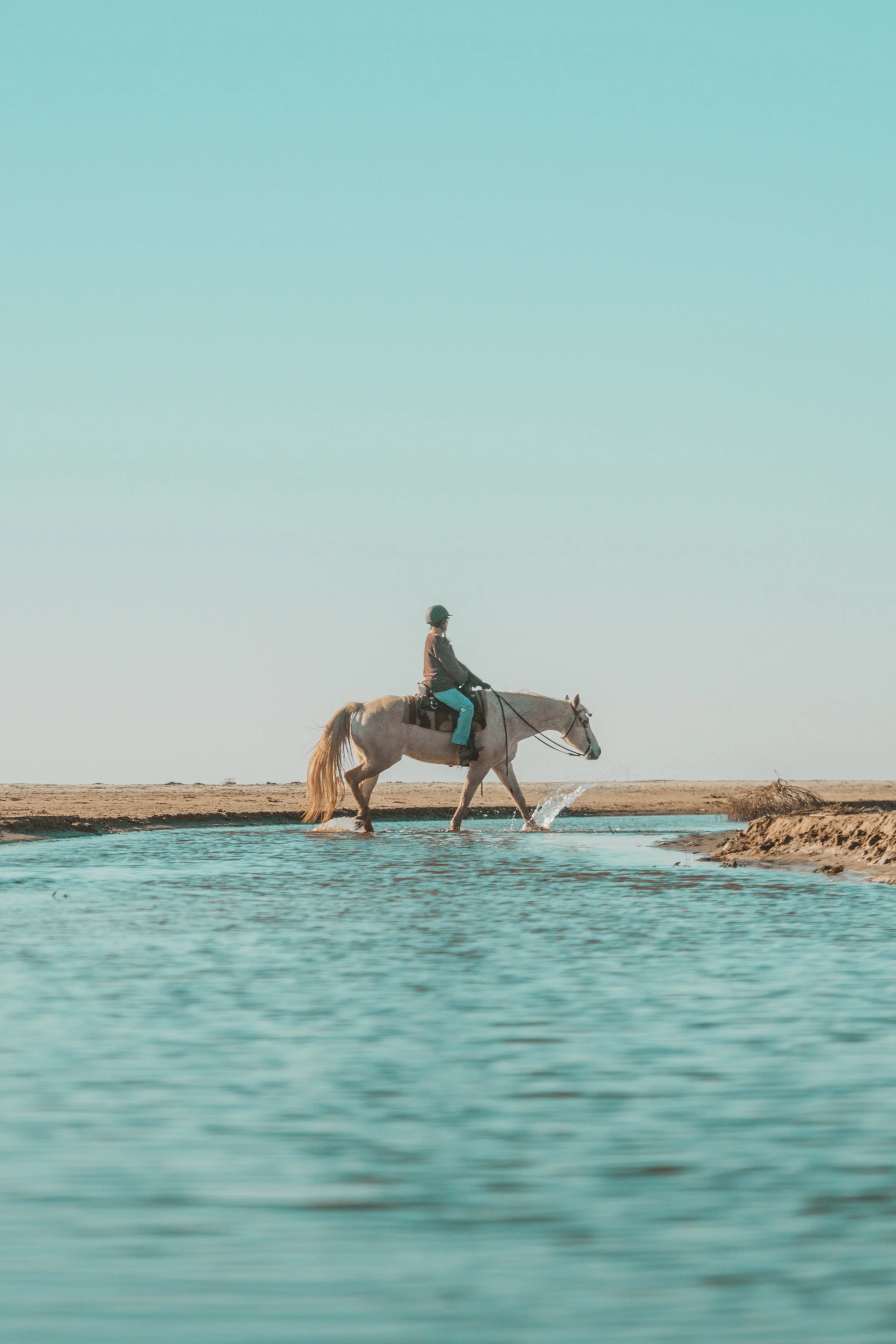 A person riding a white horse across a shallow body of water in a desert landscape under a clear blue sky.