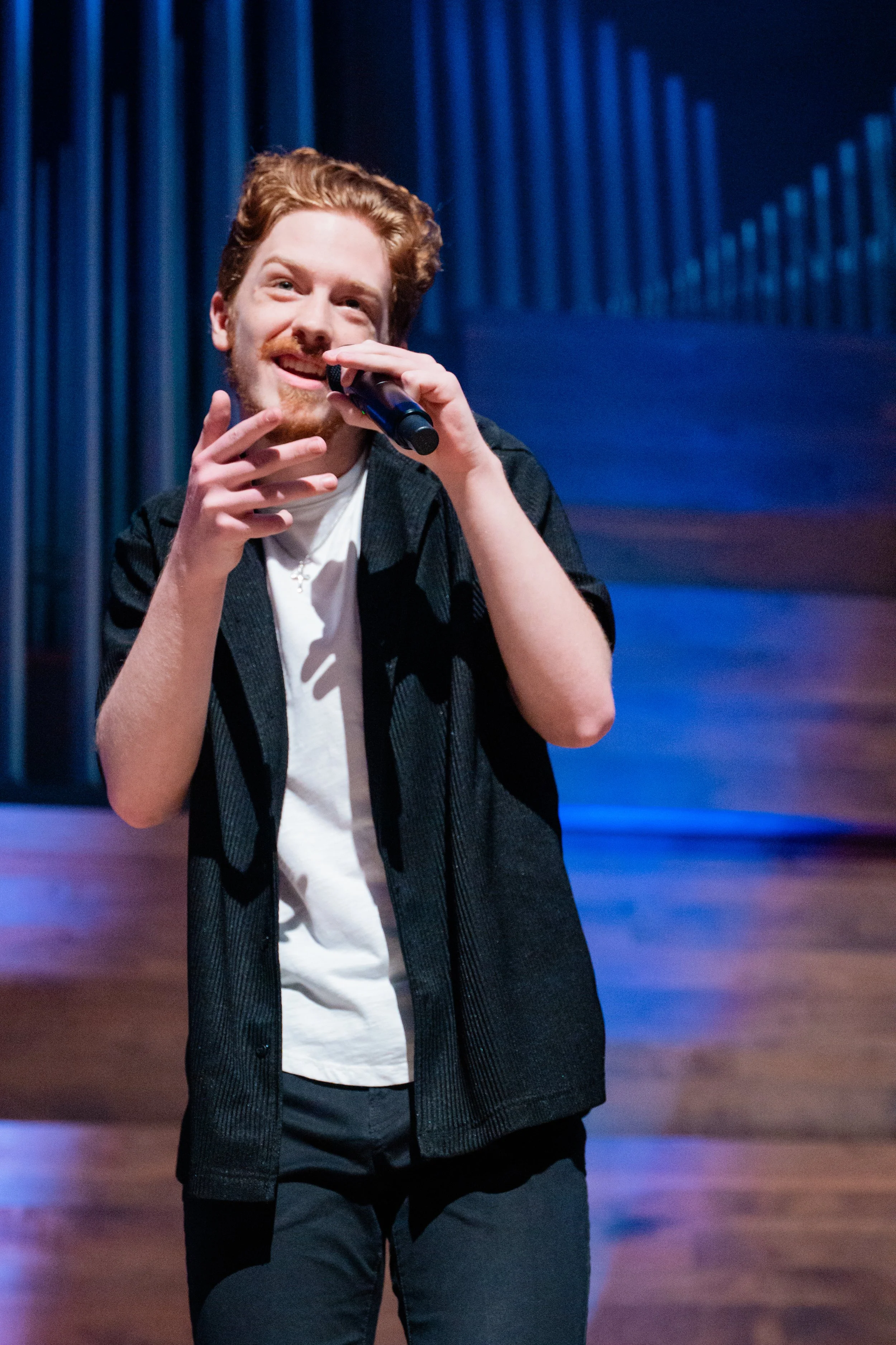 A young man with red hair and beard singing into a microphone on stage with blue and brown background.