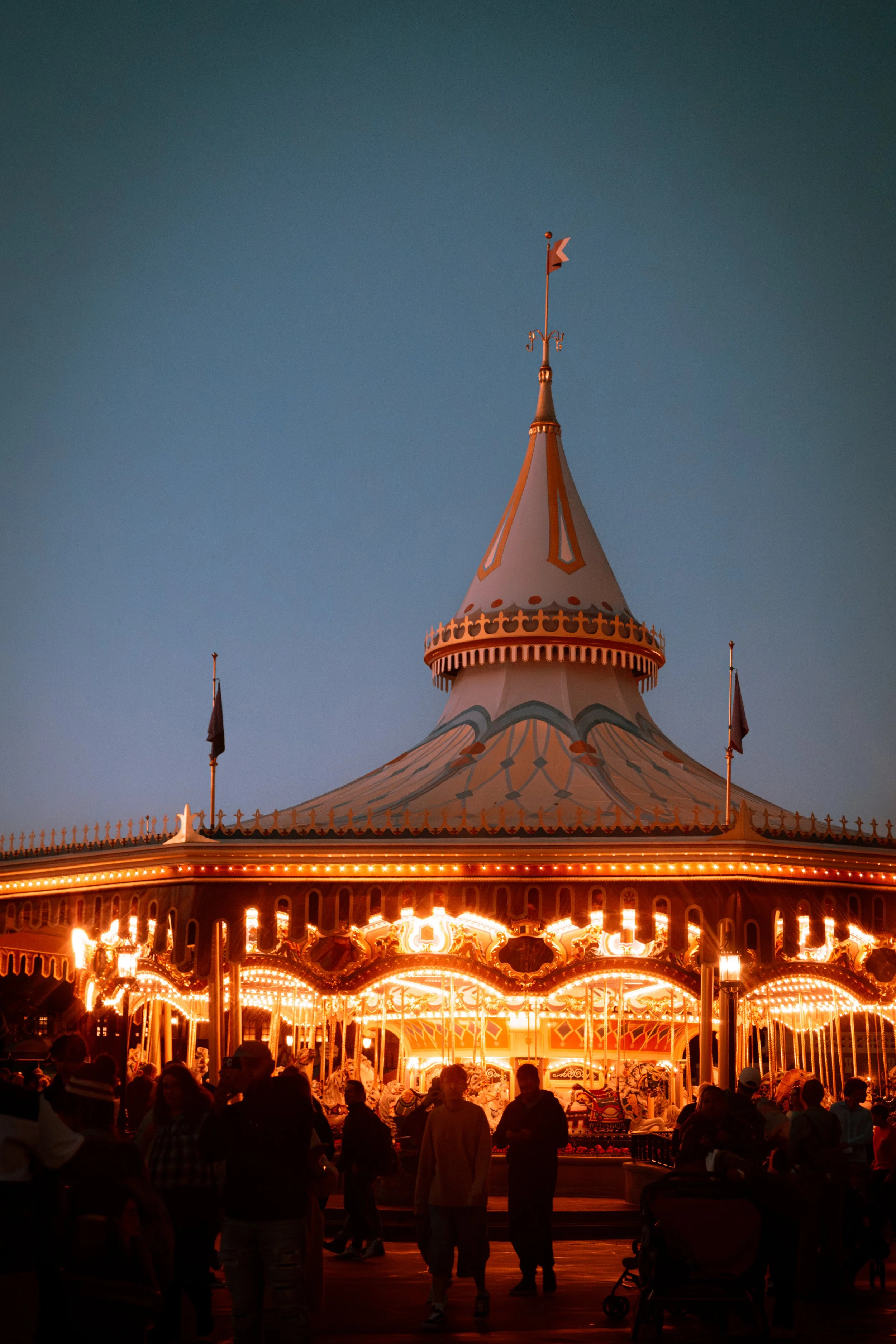 A carousel at an amusement park illuminated with bright lights during dusk, with people walking and standing around.