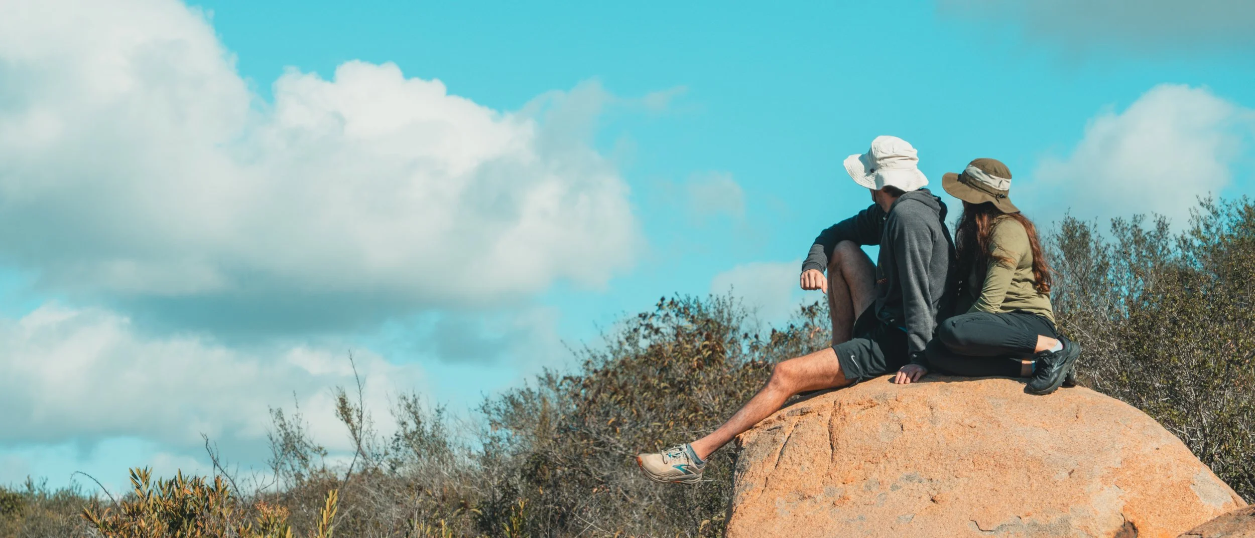 Two hikers, one male and one female, sitting on a large rock with bushy vegetation beneath a partly cloudy sky.