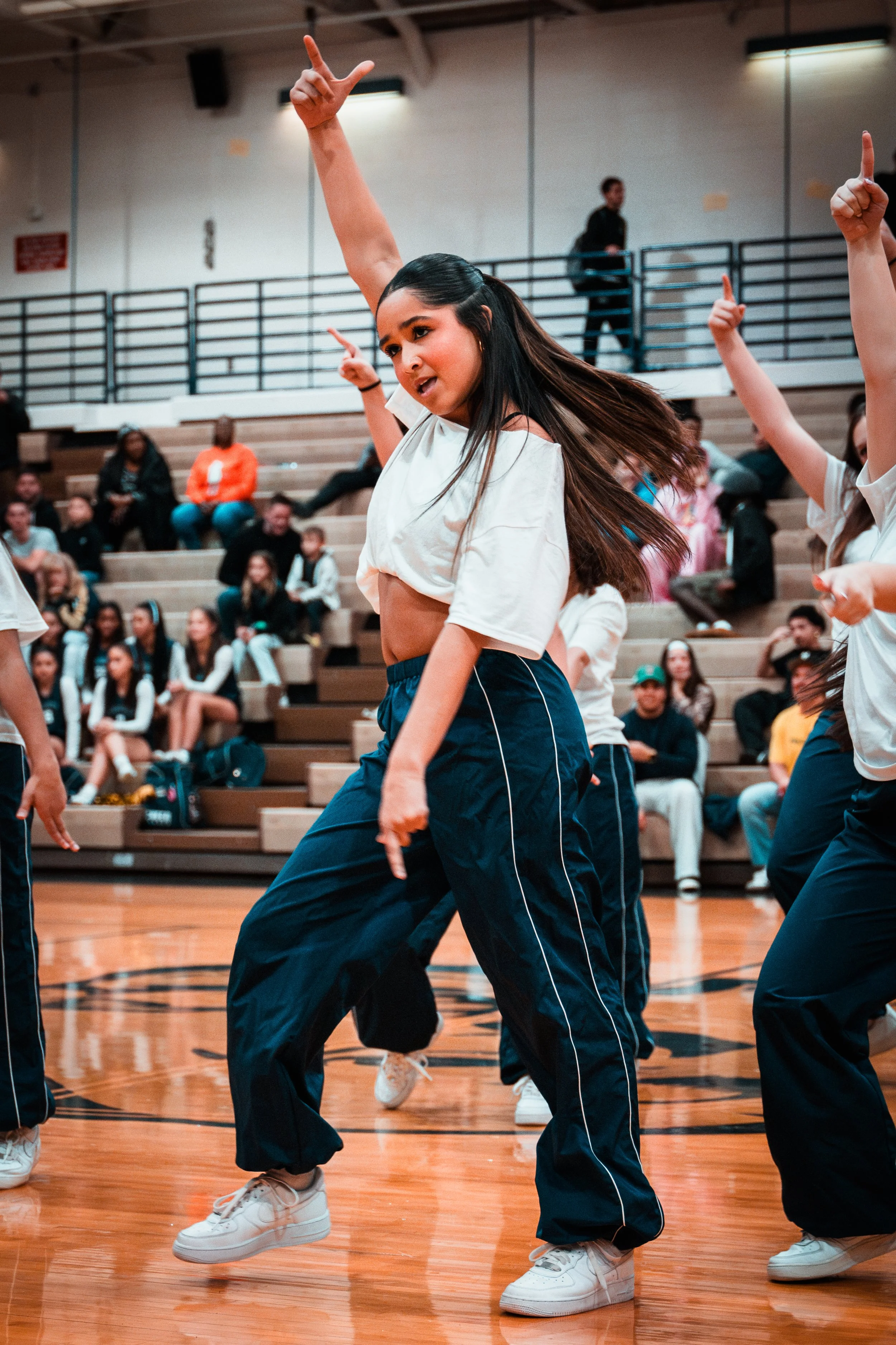 Young woman with long hair dancing in a gymnasium, surrounded by other dancers, audience seated on bleachers in the background.