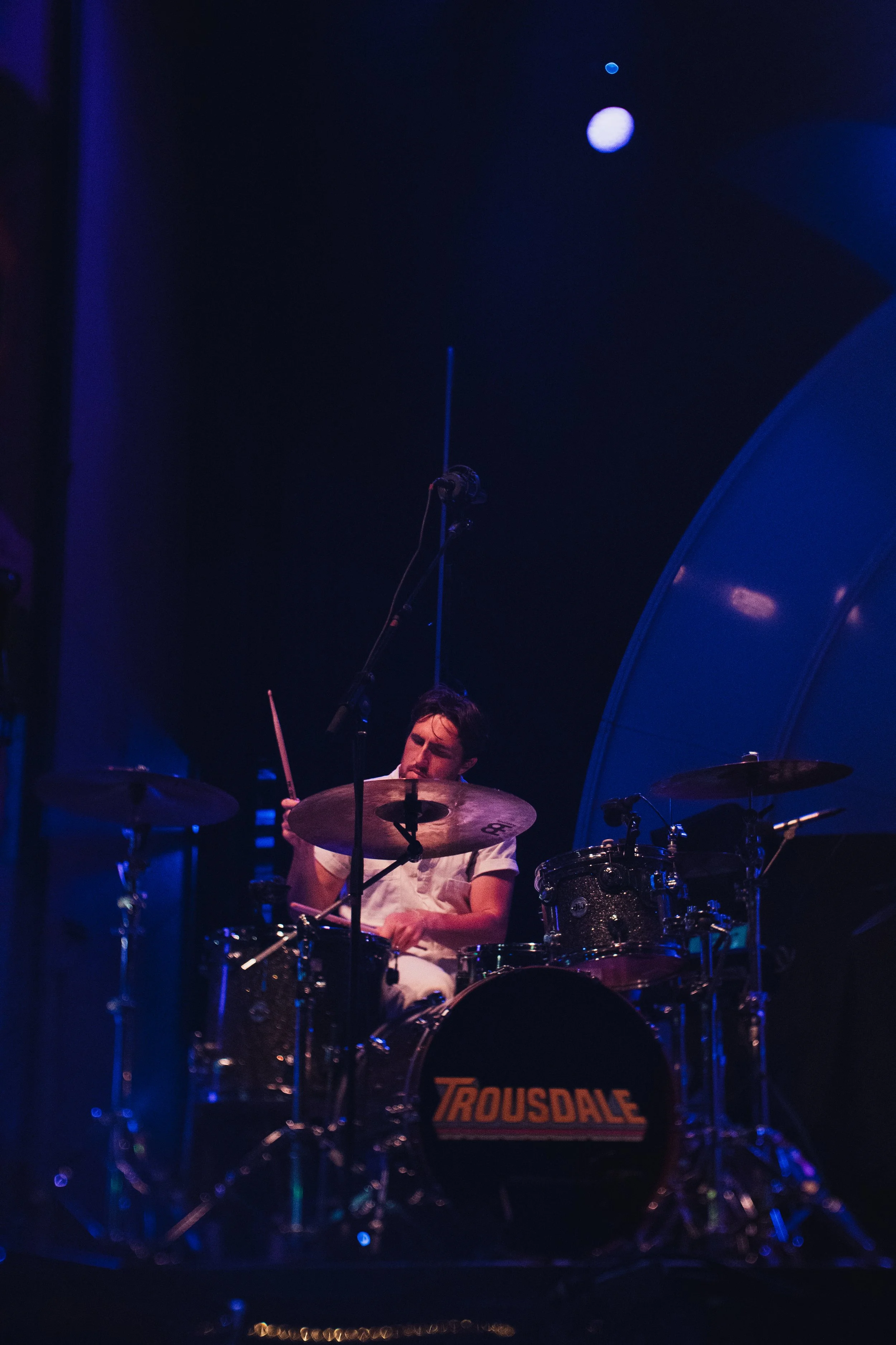 A man playing a drum set on stage, lit with blue and purple lighting, with a moon visible in the dark sky above.