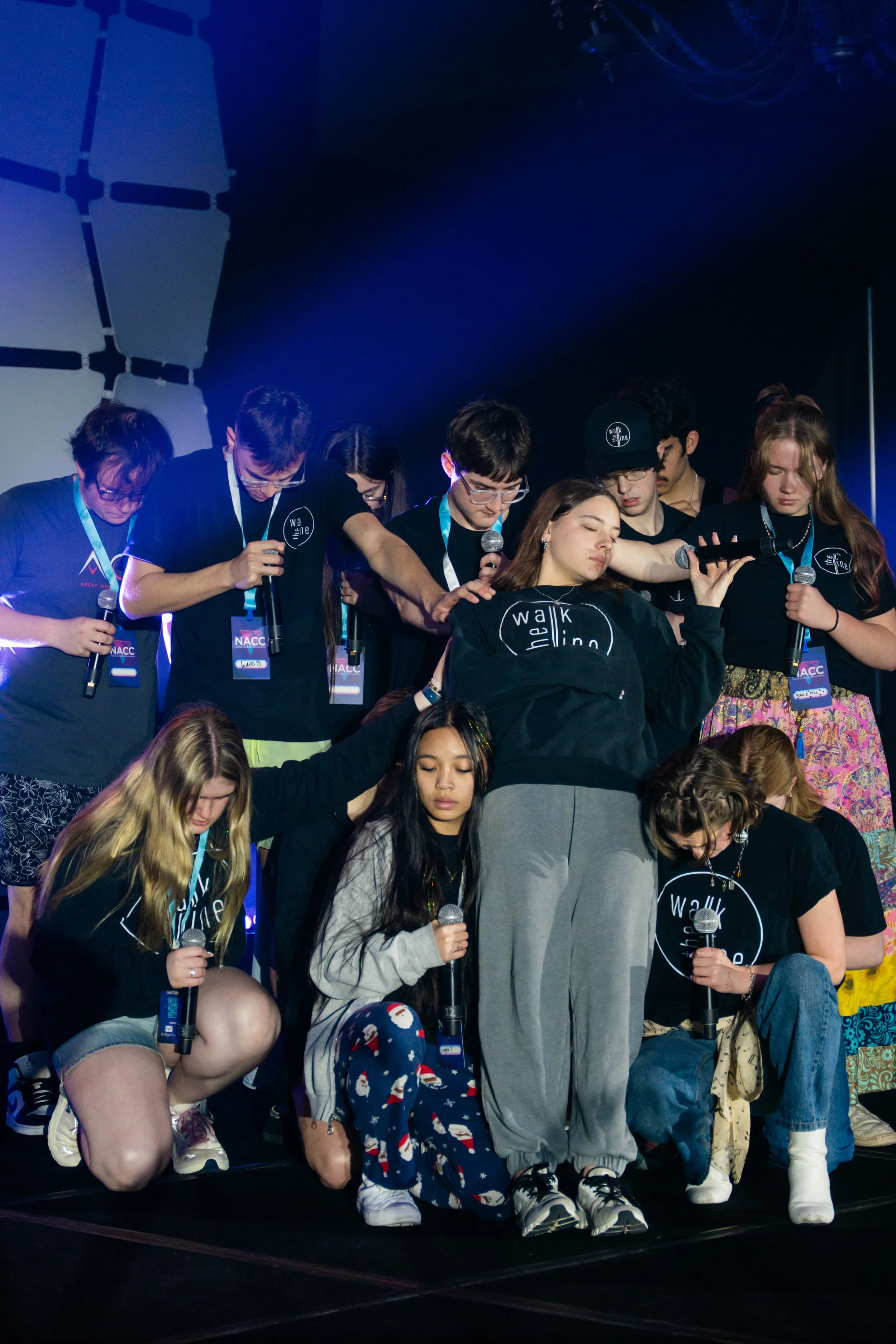 A group of young people praying or meditating with their eyes closed on a stage, some holding microphones. They are wearing casual clothes with some wearing black t-shirts with a circular logo 'walk the line'. The background is dark with stage lights