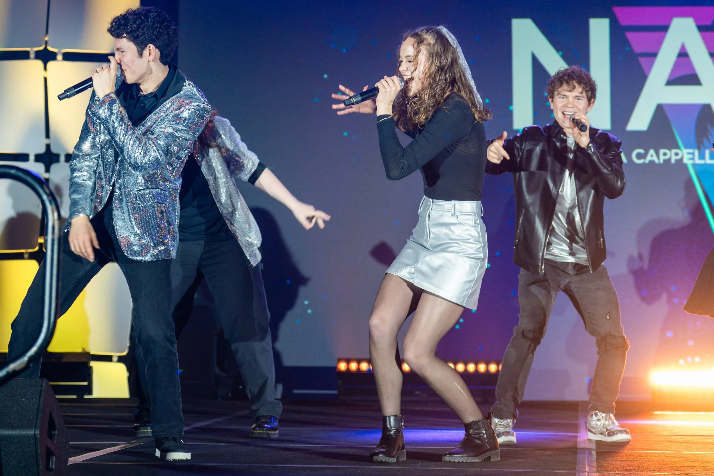 Four young performers singing into microphones on stage during a concert. The background displays a colorful light design with text.