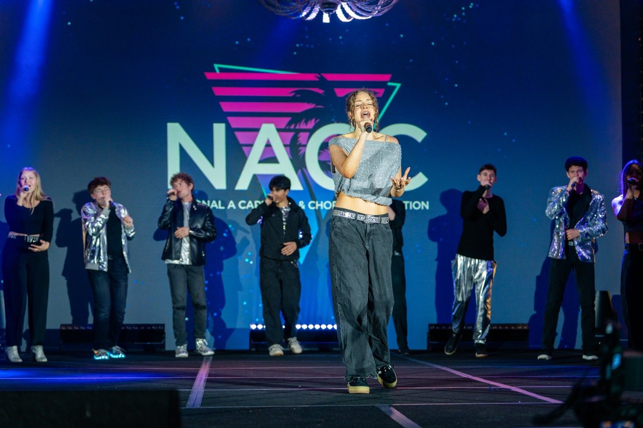 Teenagers performing on stage at NACC event, some singing with microphones, some dancing, with a large NACC logo and colorful background projection.