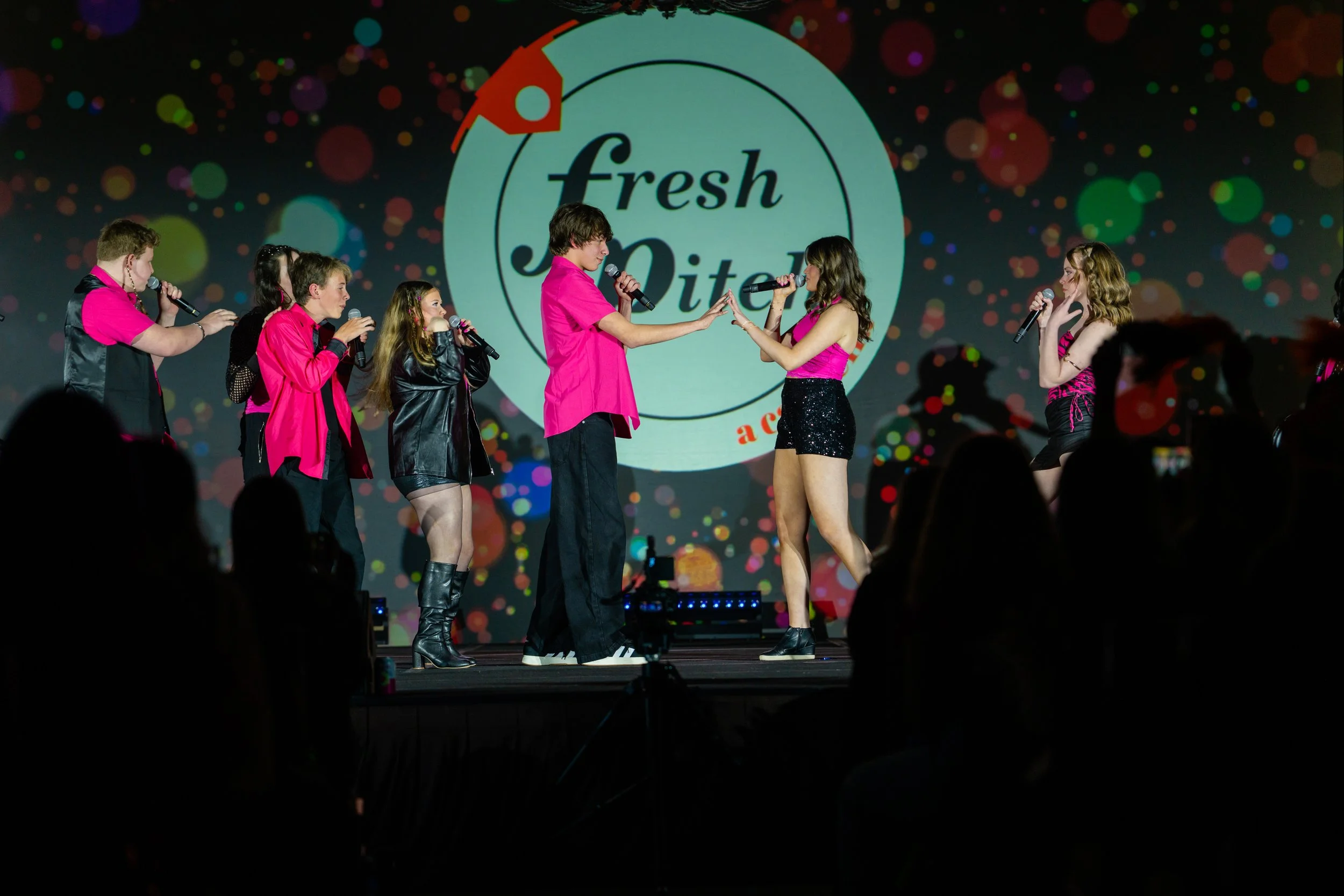 A group of young performers on stage during a talent show. The background displays a large sign that reads 'Fresh Spark' with colorful, dotted decorations. The performers are dressed in pink and black outfits, holding microphones, and engaging in a t