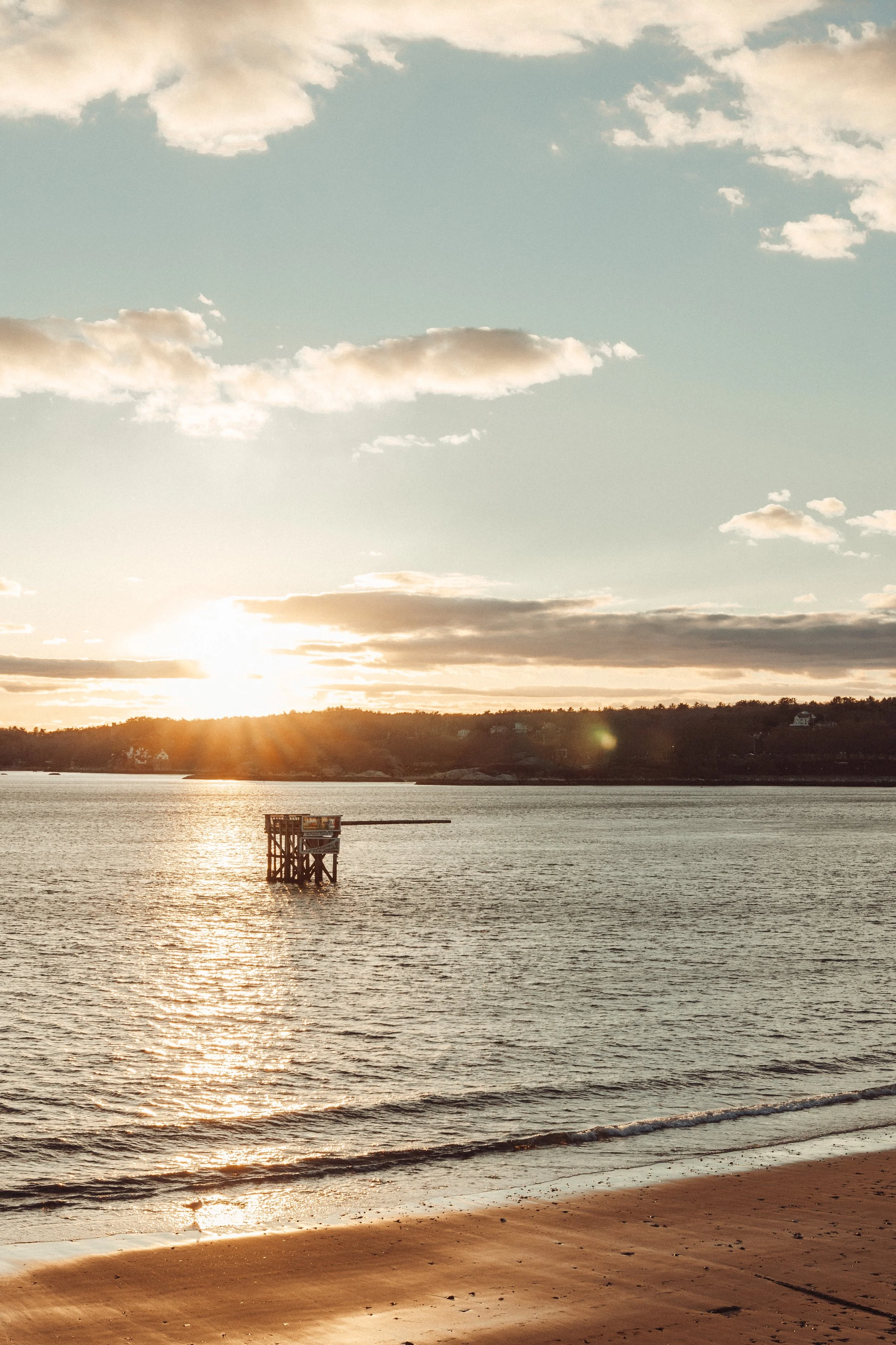 Sunset over a body of water with a wooden structure in the water and a sandy beach in the foreground.