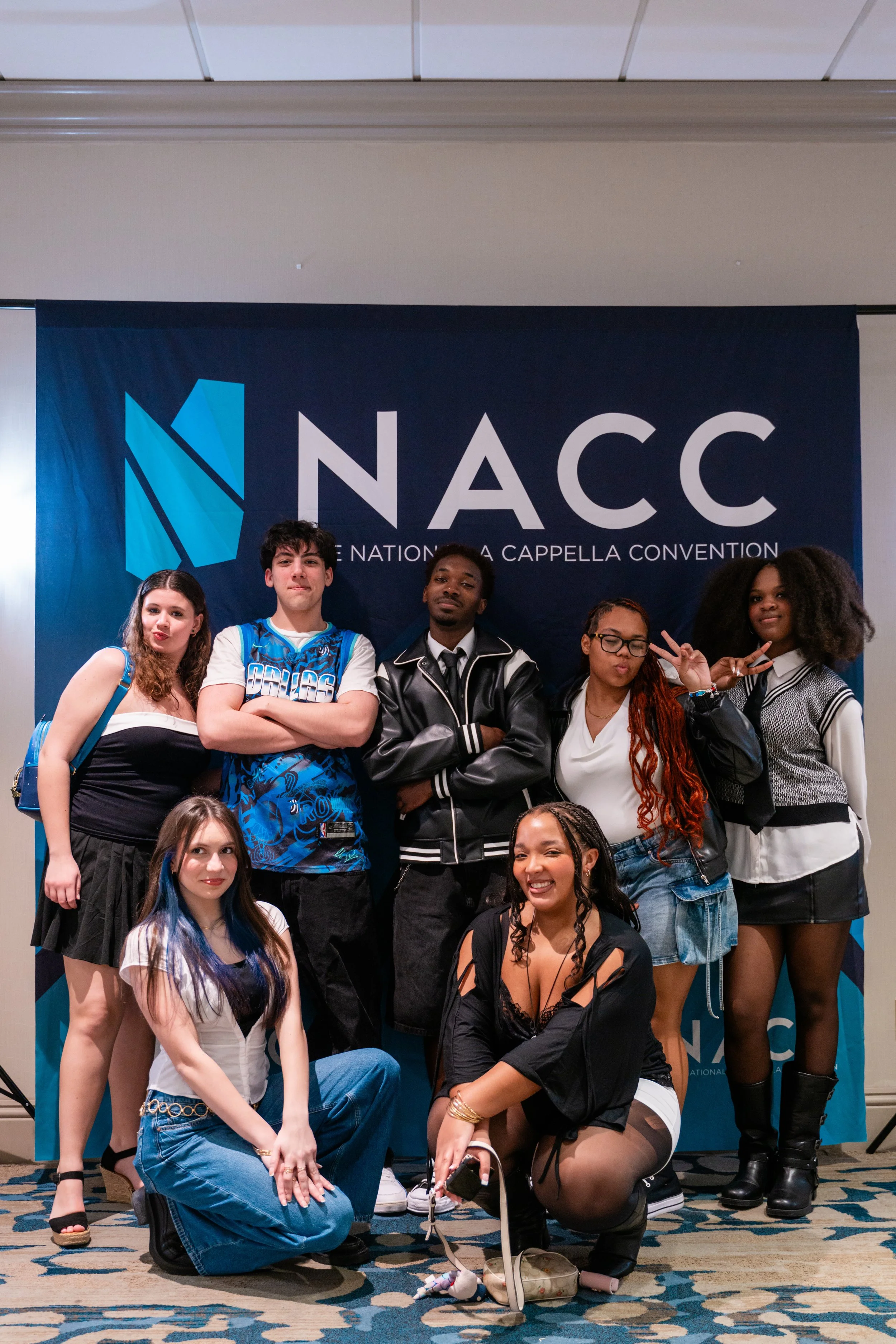A group of young diverse people posing together at the NACC National A Cappella Convention, standing in front of a large blue banner with the event's logo and name. Some are standing, some kneeling, and all are displaying different expressions and ge