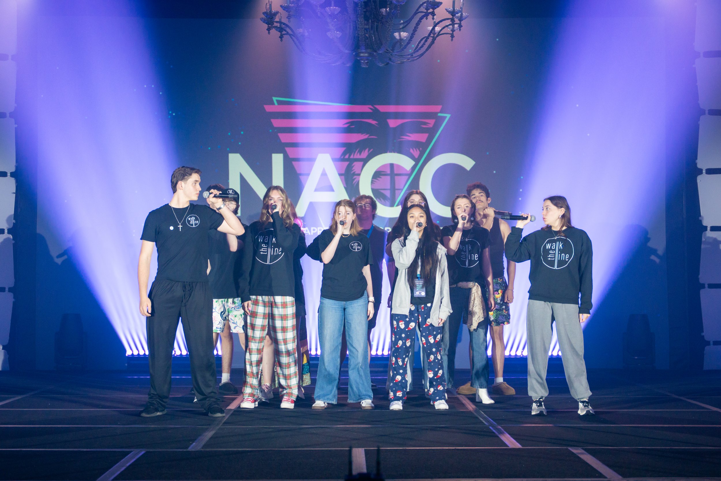 Group of diverse teenagers standing on stage with a neon NASA logo in the background, some holding microphones, dressed in casual clothing with some wearing pajamas and sportswear.