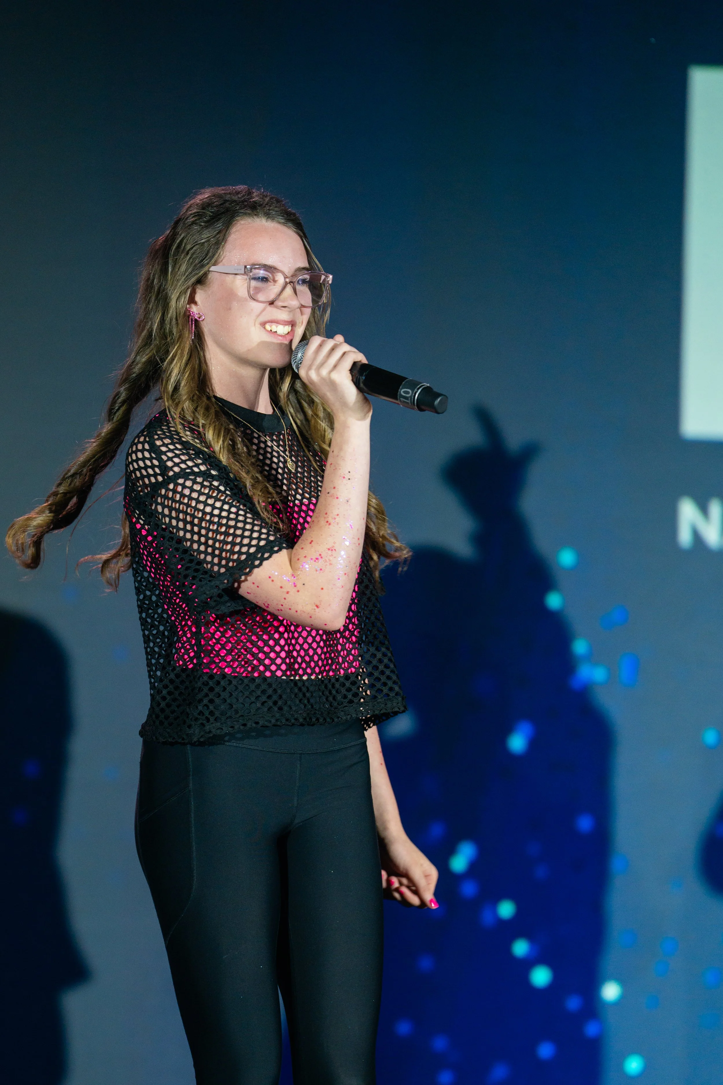 A young woman with glasses and long wavy hair holding a microphone, wearing a black fishnet top with pink underneath and black pants, standing on stage with a blue background and blurry lights.