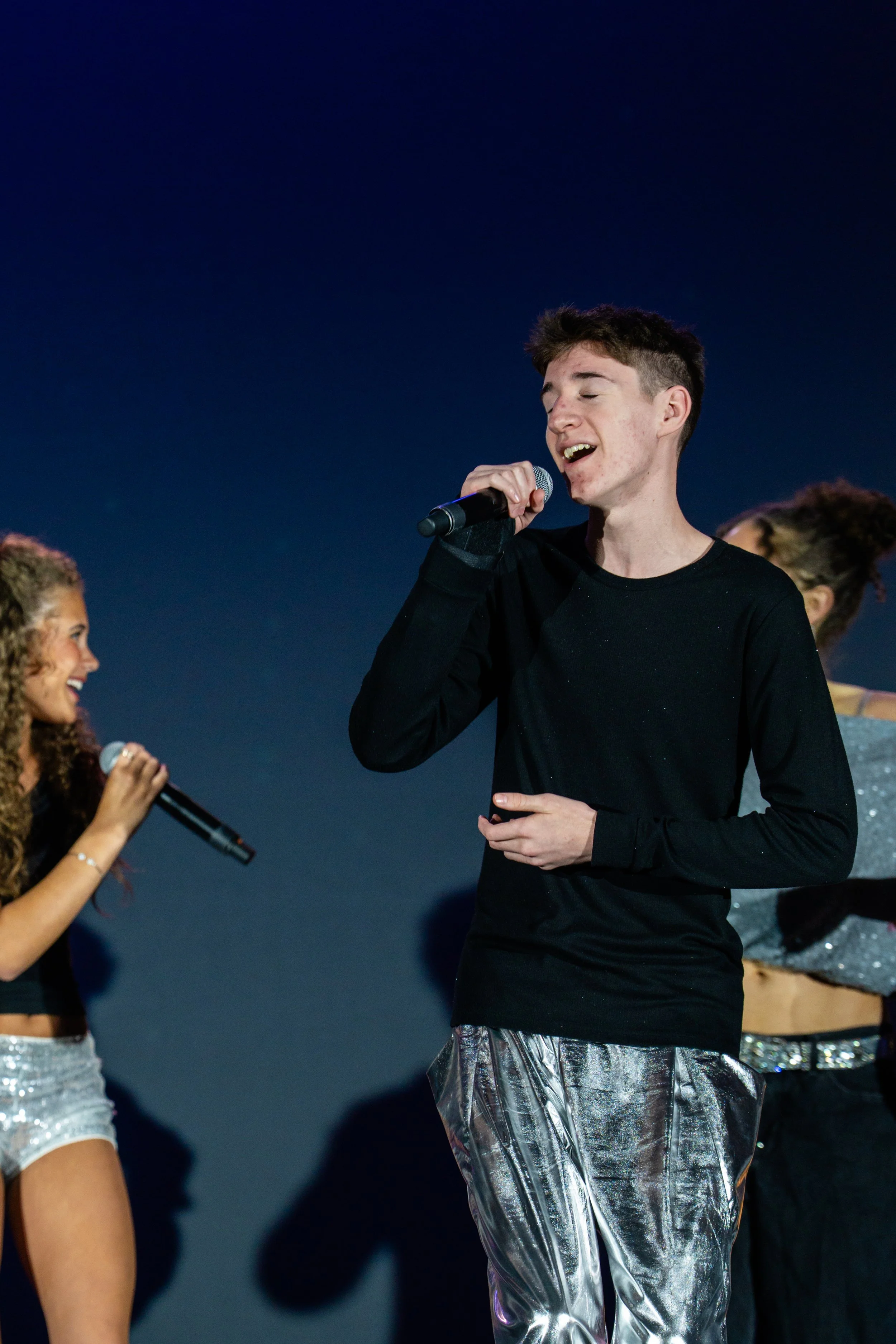 Young man singing into a microphone on stage with a dark blue background, surrounded by backup singers with microphones and metallic silver pants.