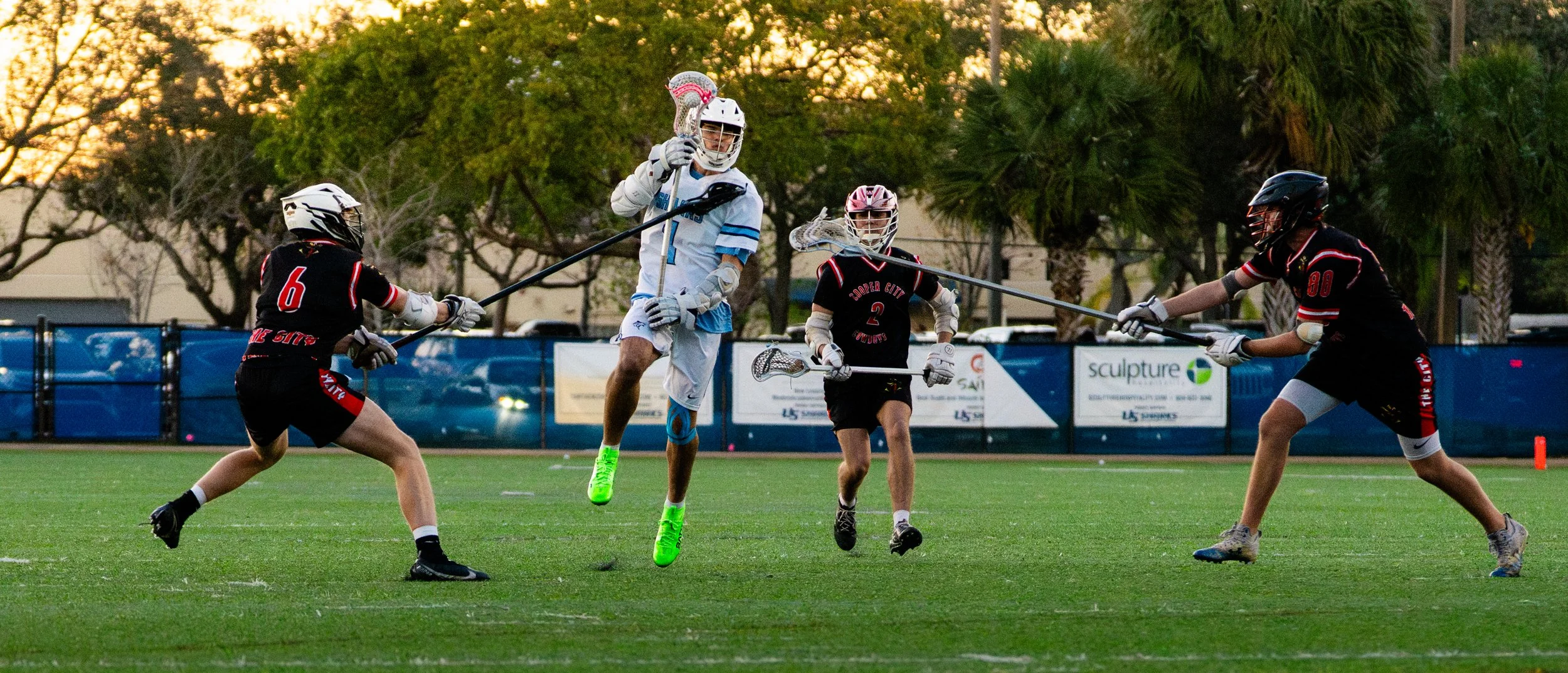 Lacrosse players in action on the field during a game at sunset, with some players wearing black and red jerseys and a player in a white and blue uniform jumping.