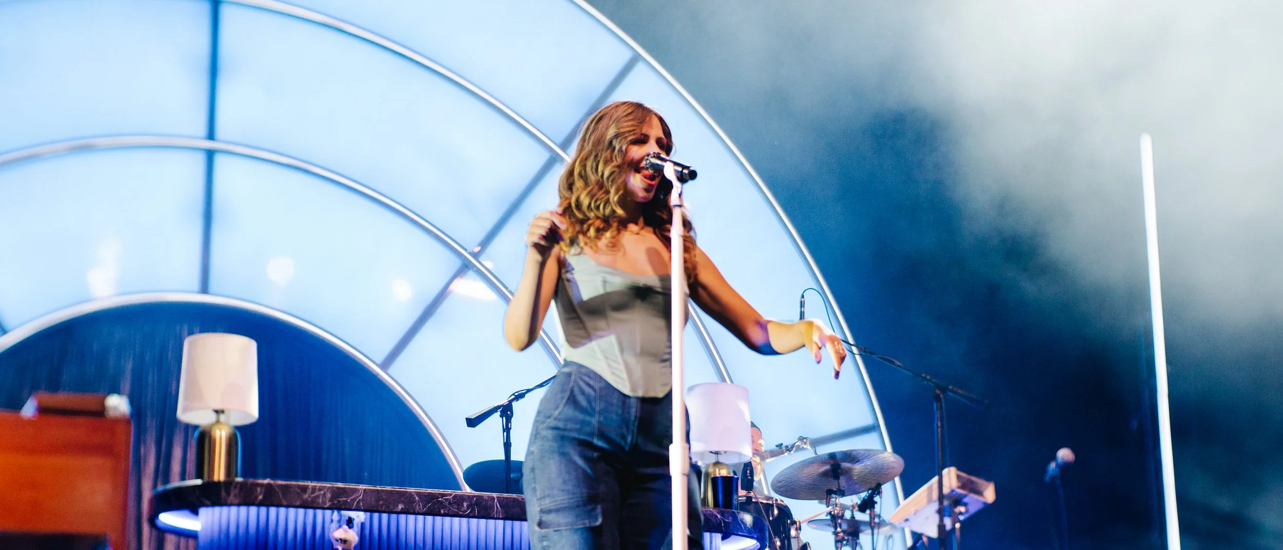 A woman singing on stage with a microphone in front of her, with stage lighting and musical instruments around her.