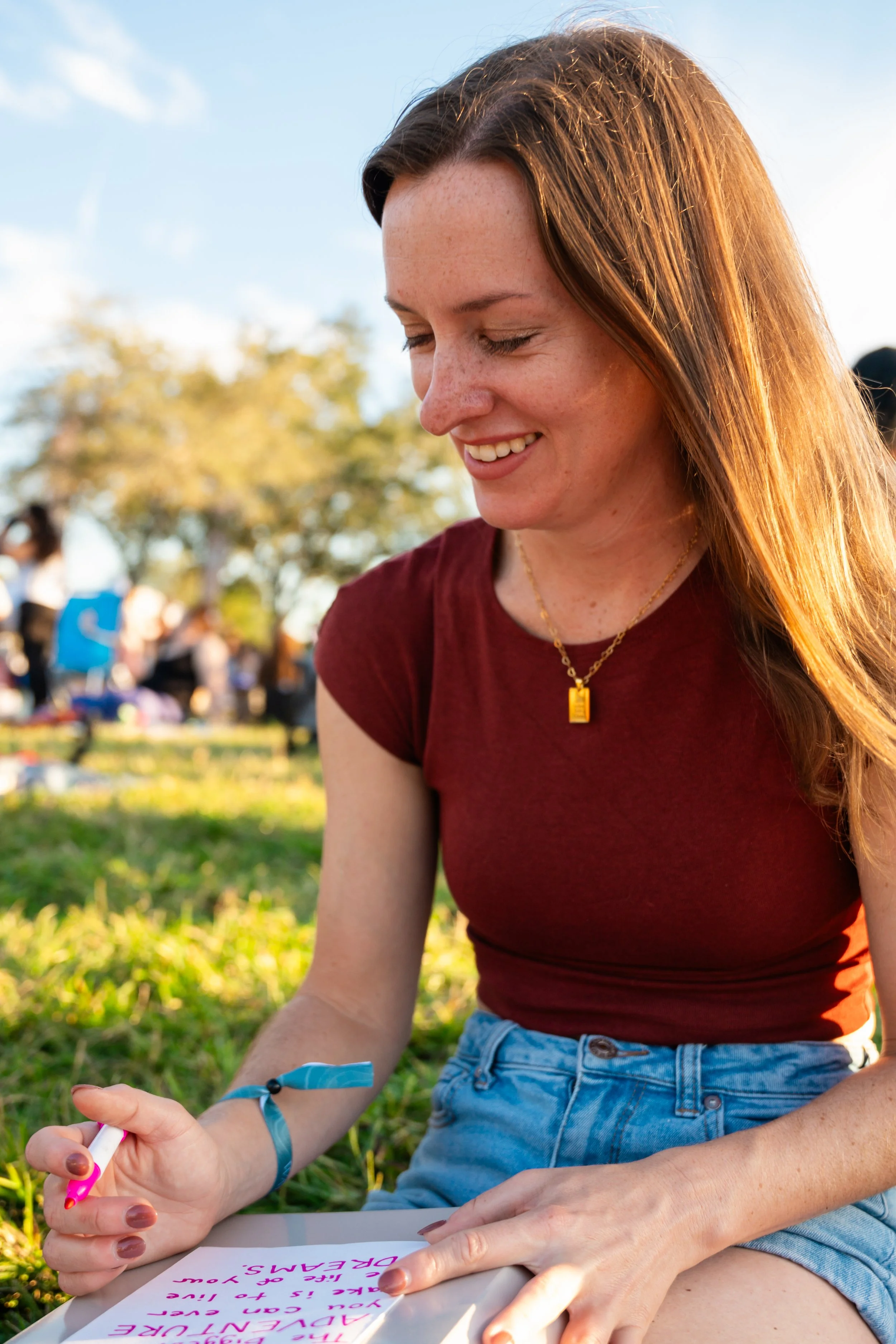 A young woman with long red hair is sitting outdoors on grass, smiling and writing on a piece of paper with a pink pen. She wears a maroon t-shirt, high-waisted denim shorts, a gold necklace, and a blue wristband. In the background, there are other p