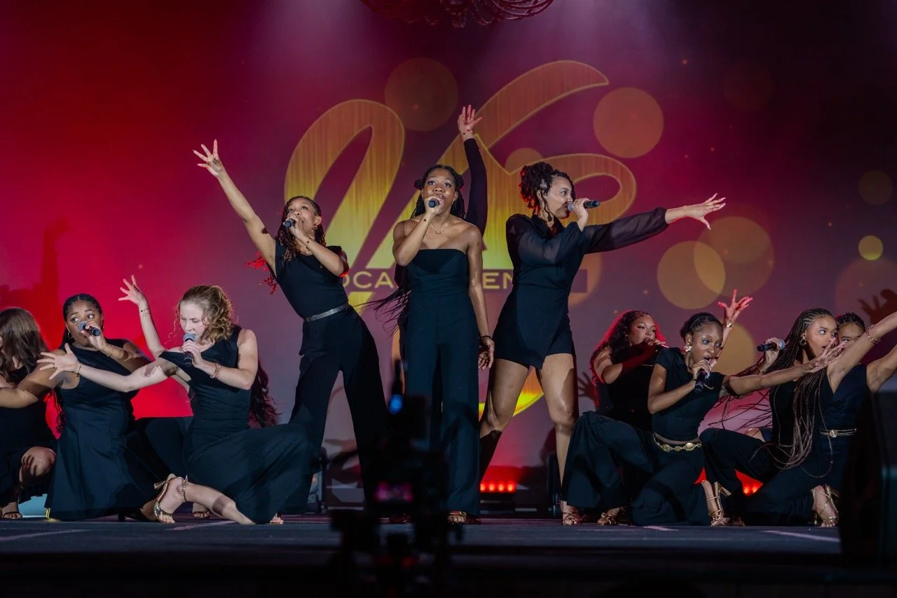 Group of ten diverse women performing on stage with microphones, dressed in black, with a large logo in the background.