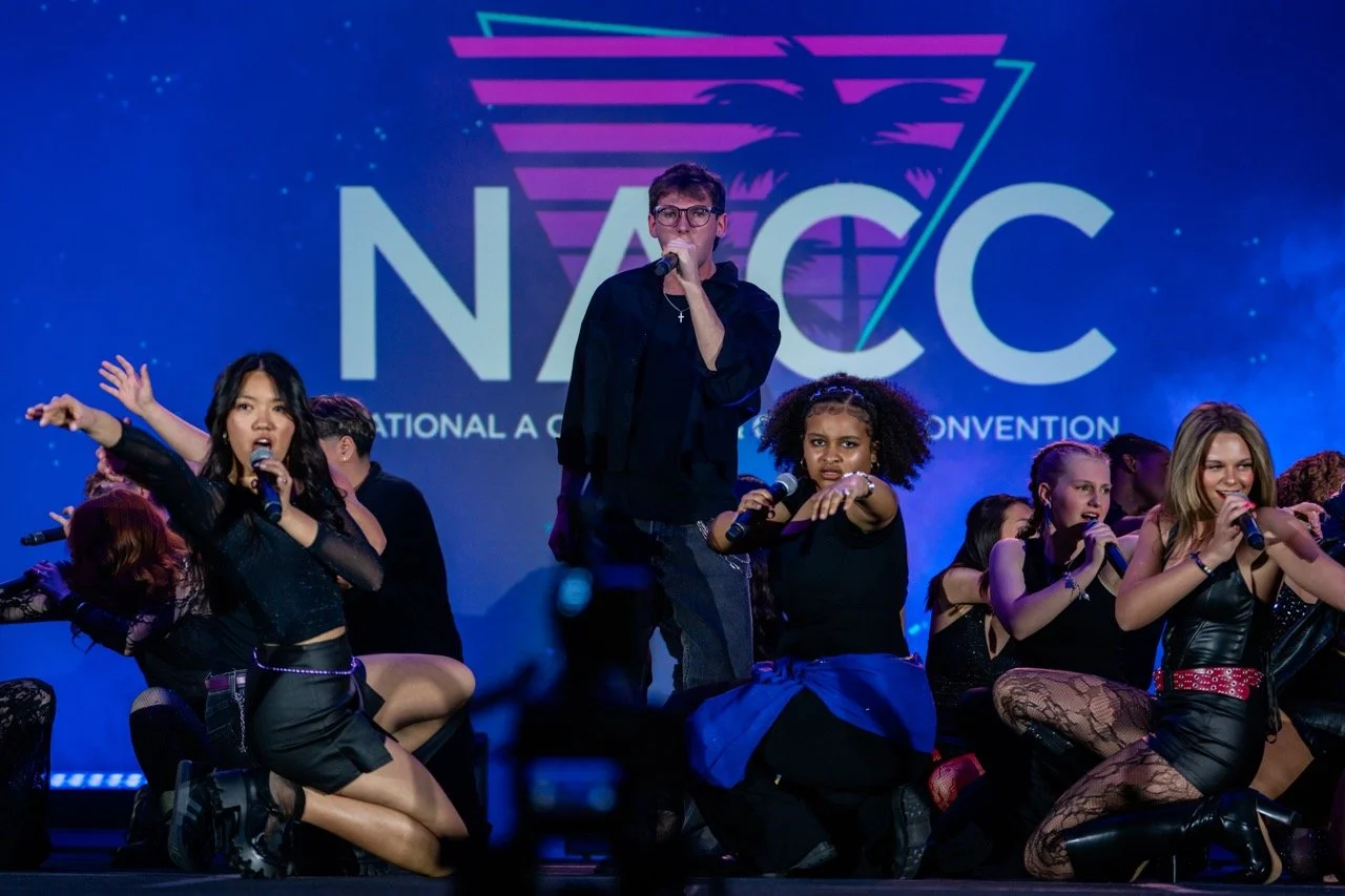 A group of young performers on stage at the National A Cappella Convention with a large NACC logo in the background.