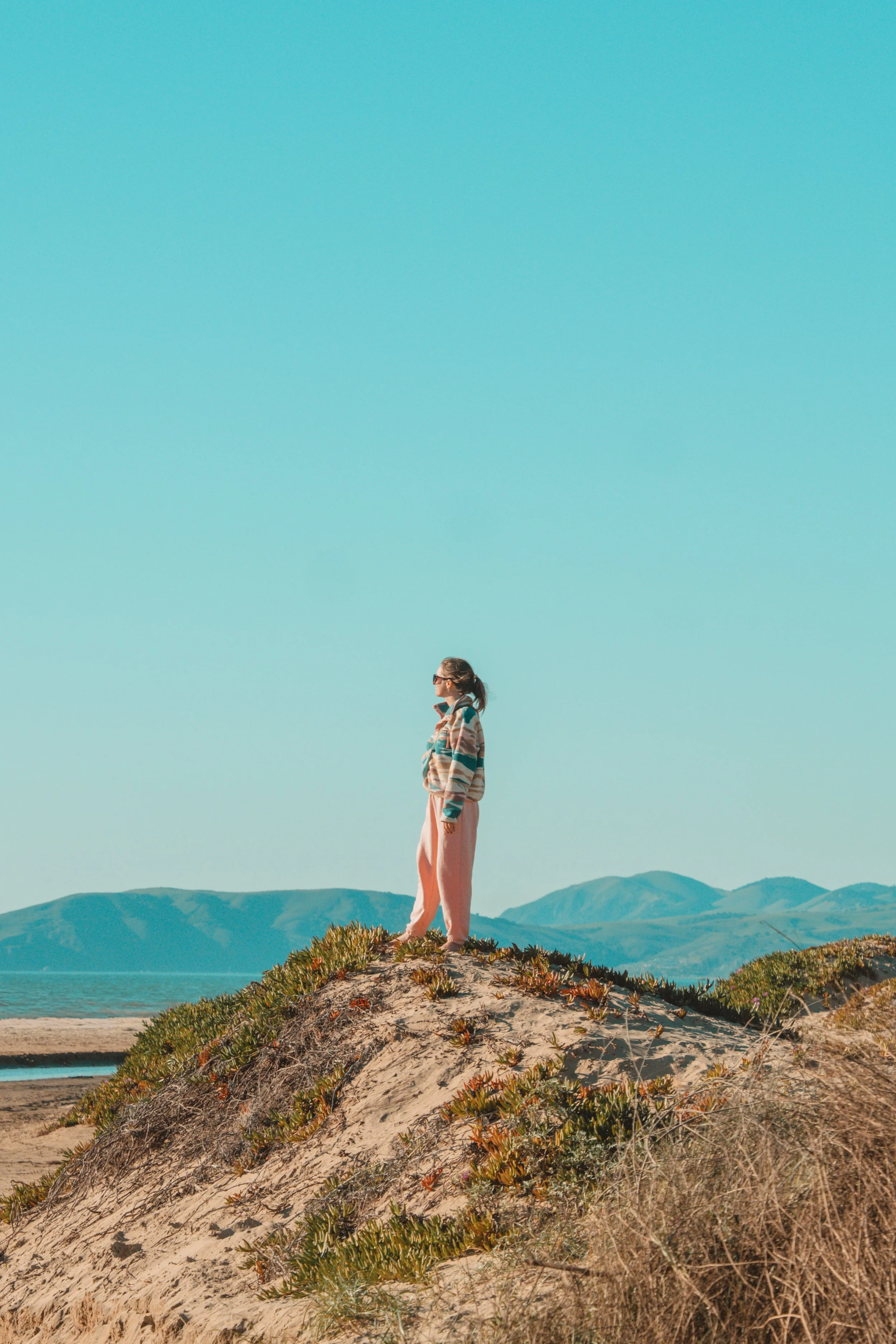 A woman standing on a sandy hill with green plants, overlooking the ocean and mountains in the distance, under a clear blue sky.